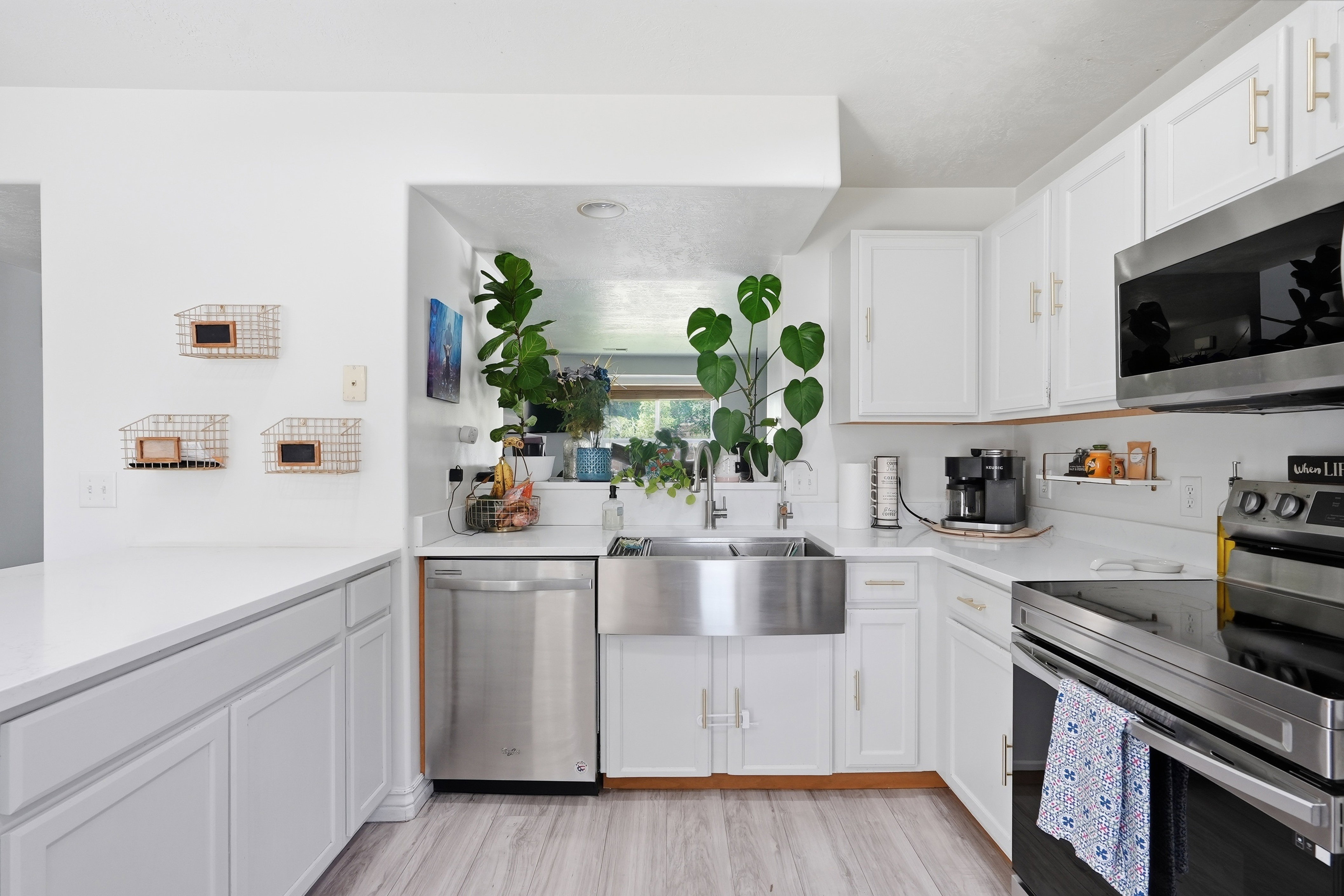 White kitchen cabinetry with gold-tone hardware, stainless steel farmhouse sink, stainless steel dishwasher, stainless steel microwave, and wood-finish flooring
