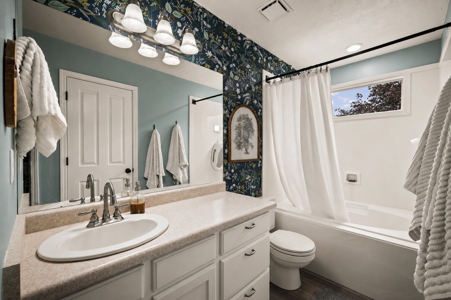 Bathroom featuring an oval undermount sink, a light-toned countertop, a full-width vanity mirror, a bathtub with a shower kit, and a window