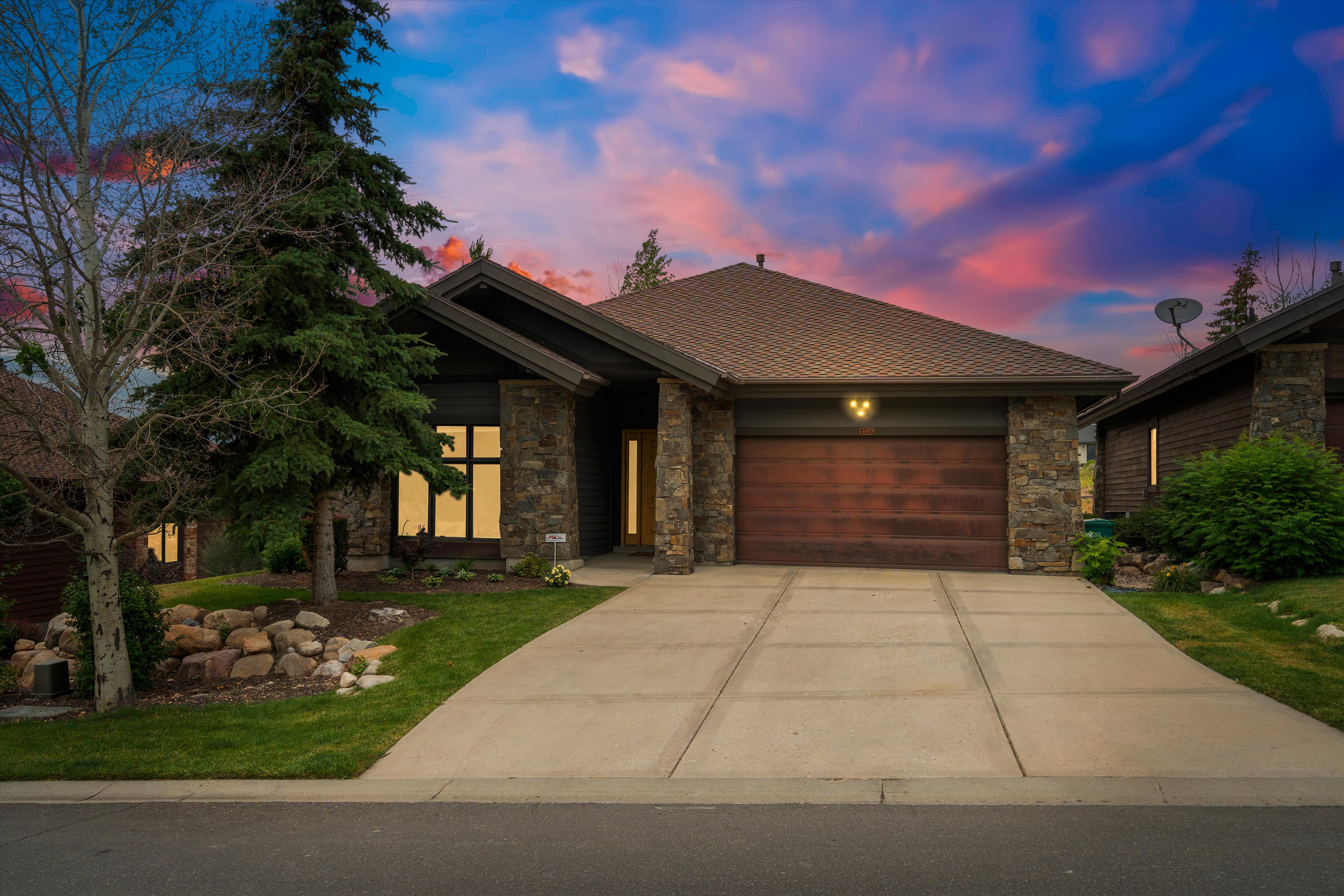Stone and wood exterior featuring a peaked roofline, an attached garage with a wood-finish door, a concrete driveway, and established landscaping with mature trees and rock elements
