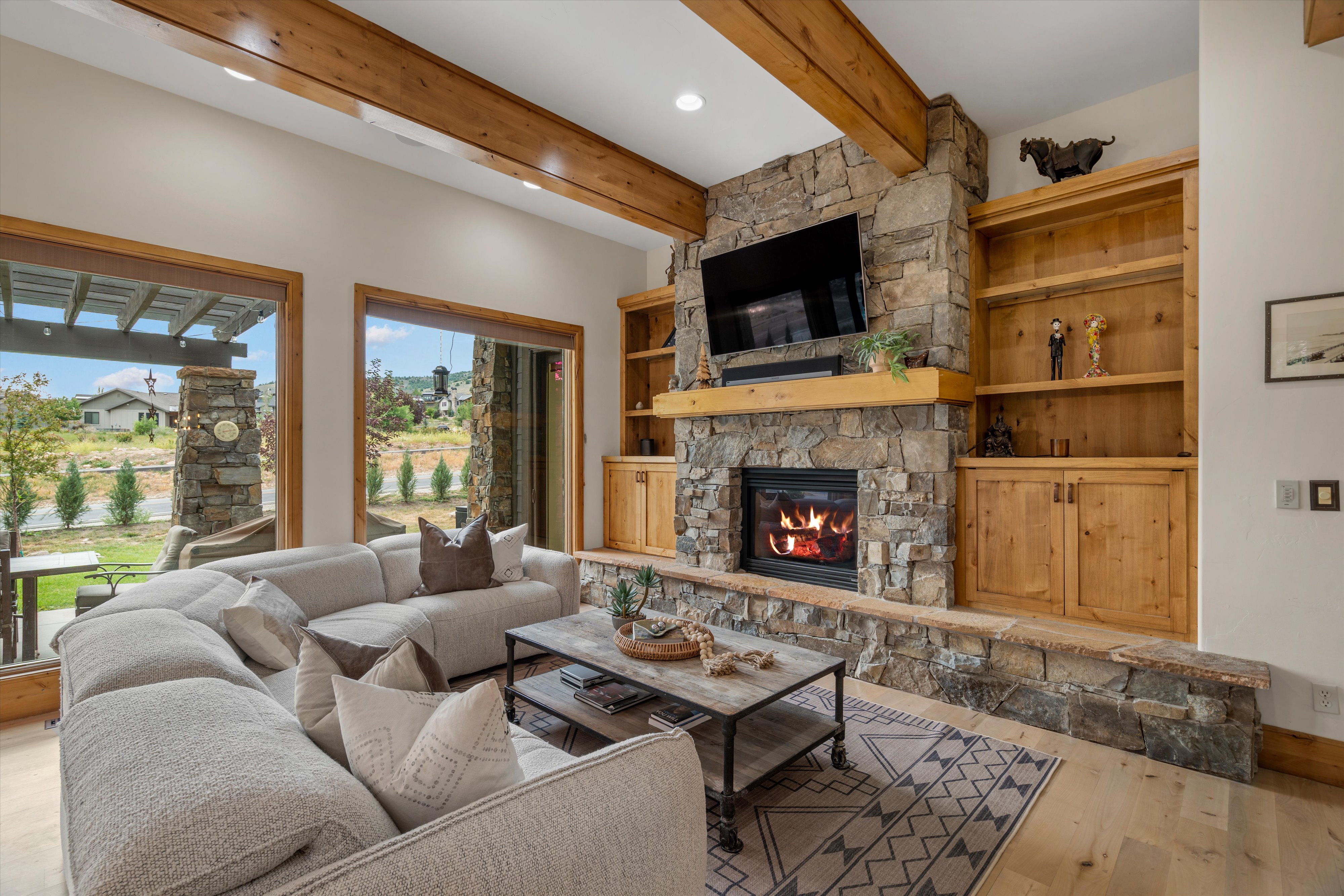 Living area featuring a stone fireplace with a wood mantle, built-in shelving, wood-finish flooring, exposed wood beams, and expansive windows