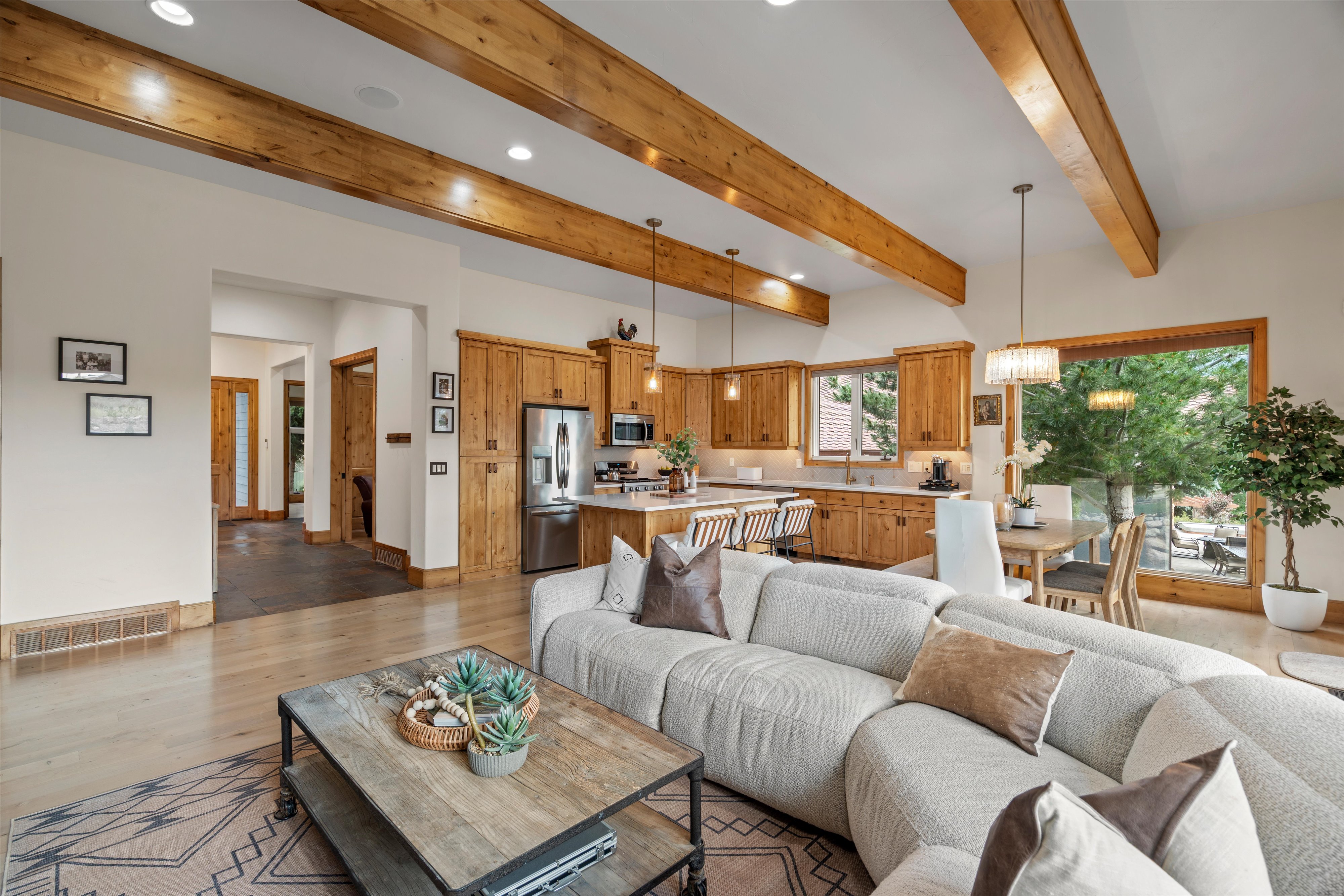 Open-concept living space featuring vaulted ceilings with exposed wood beams, light wood-finish flooring, a contemporary kitchen with wood cabinetry and stainless steel appliances, a dining area with a modern chandelier, and large windows offering exterio