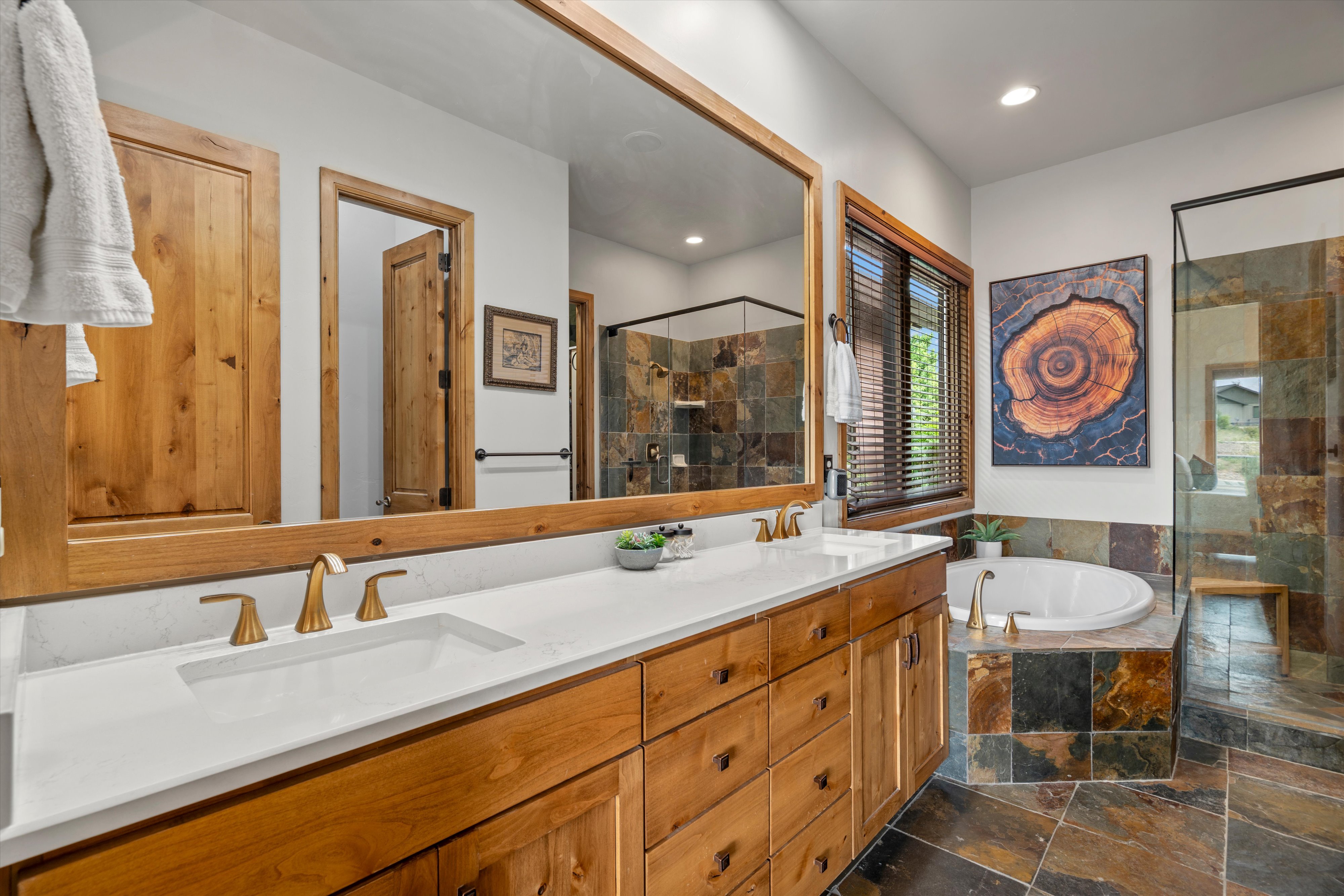 Bathroom featuring a dual vanity with white countertops and gold-finish fixtures, a large soaking tub, and an enclosed shower with stone-tile surround