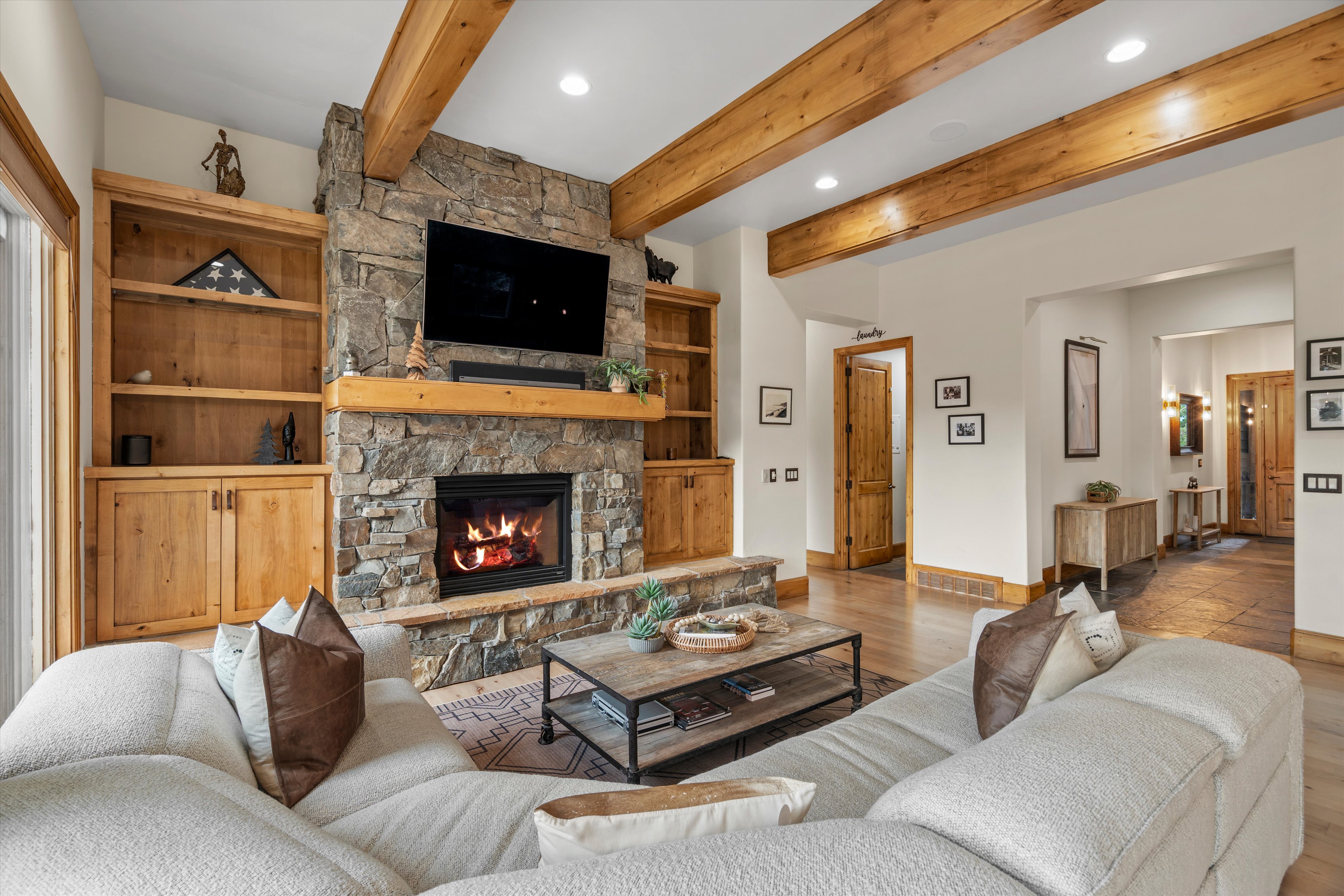 Living area featuring a stone fireplace with a wood mantle, built-in wood shelving, exposed wood ceiling beams, wood-finish flooring, and recessed lighting