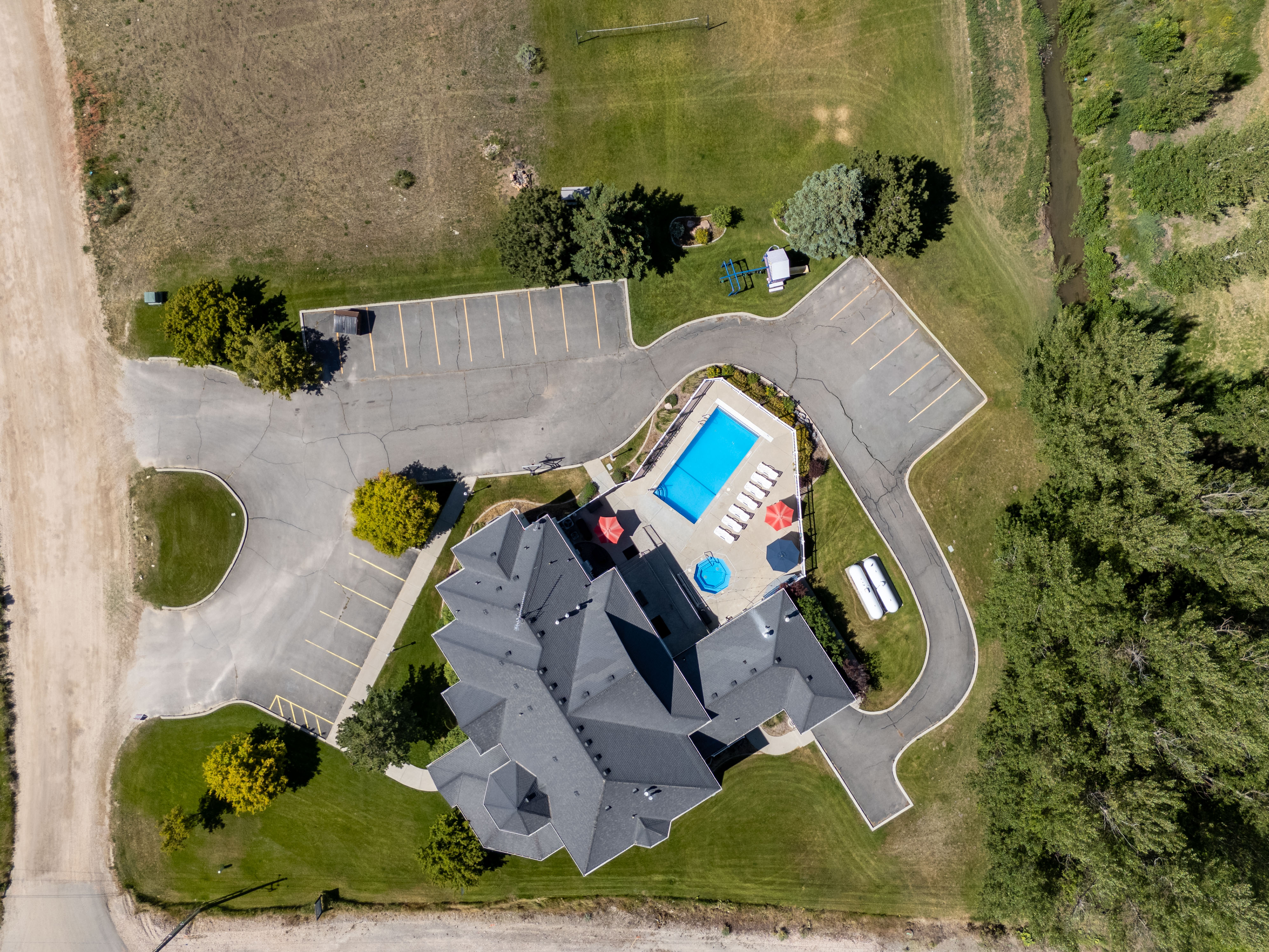 Aerial view showcasing a large gray-roofed structure, an outdoor swimming pool with surrounding patio, and extensive paved parking areas