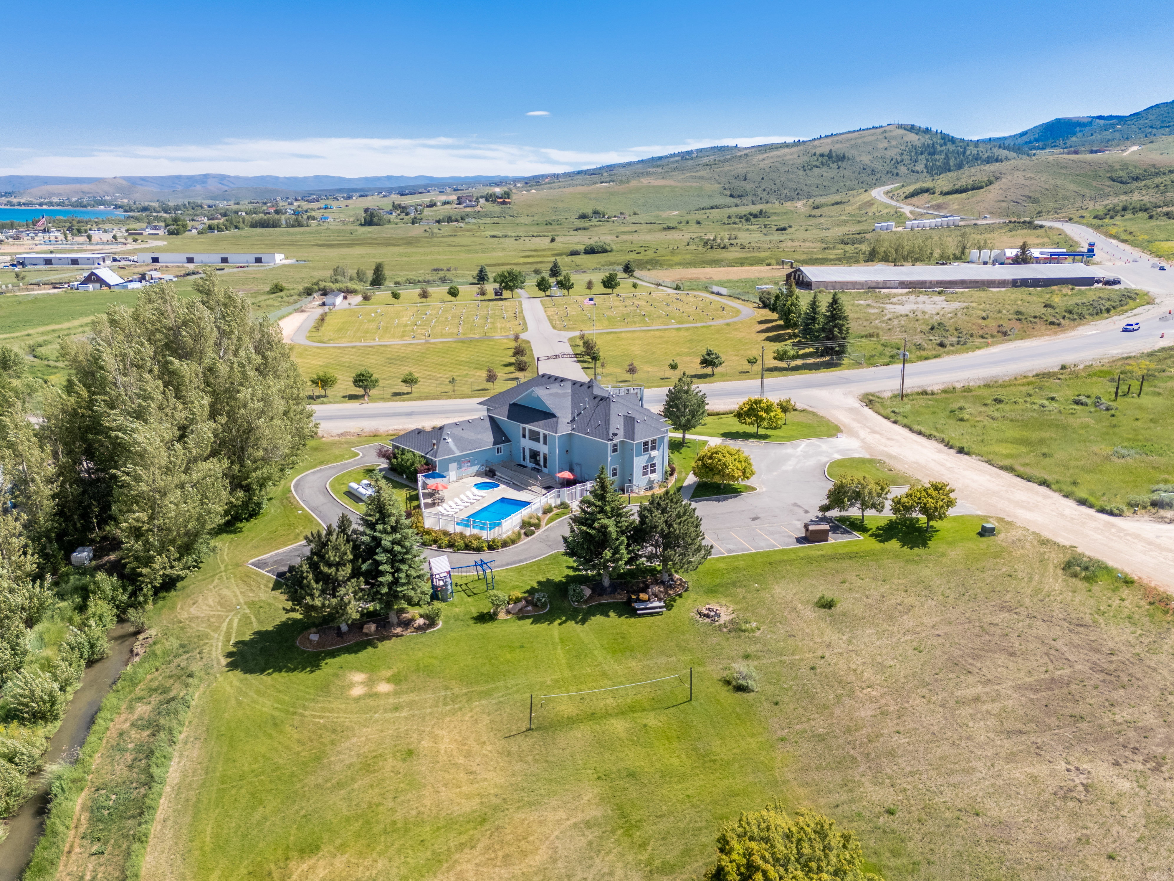 Light blue multi-story residence featuring a dark roof, a swimming pool with a surrounding patio, and mature evergreen trees