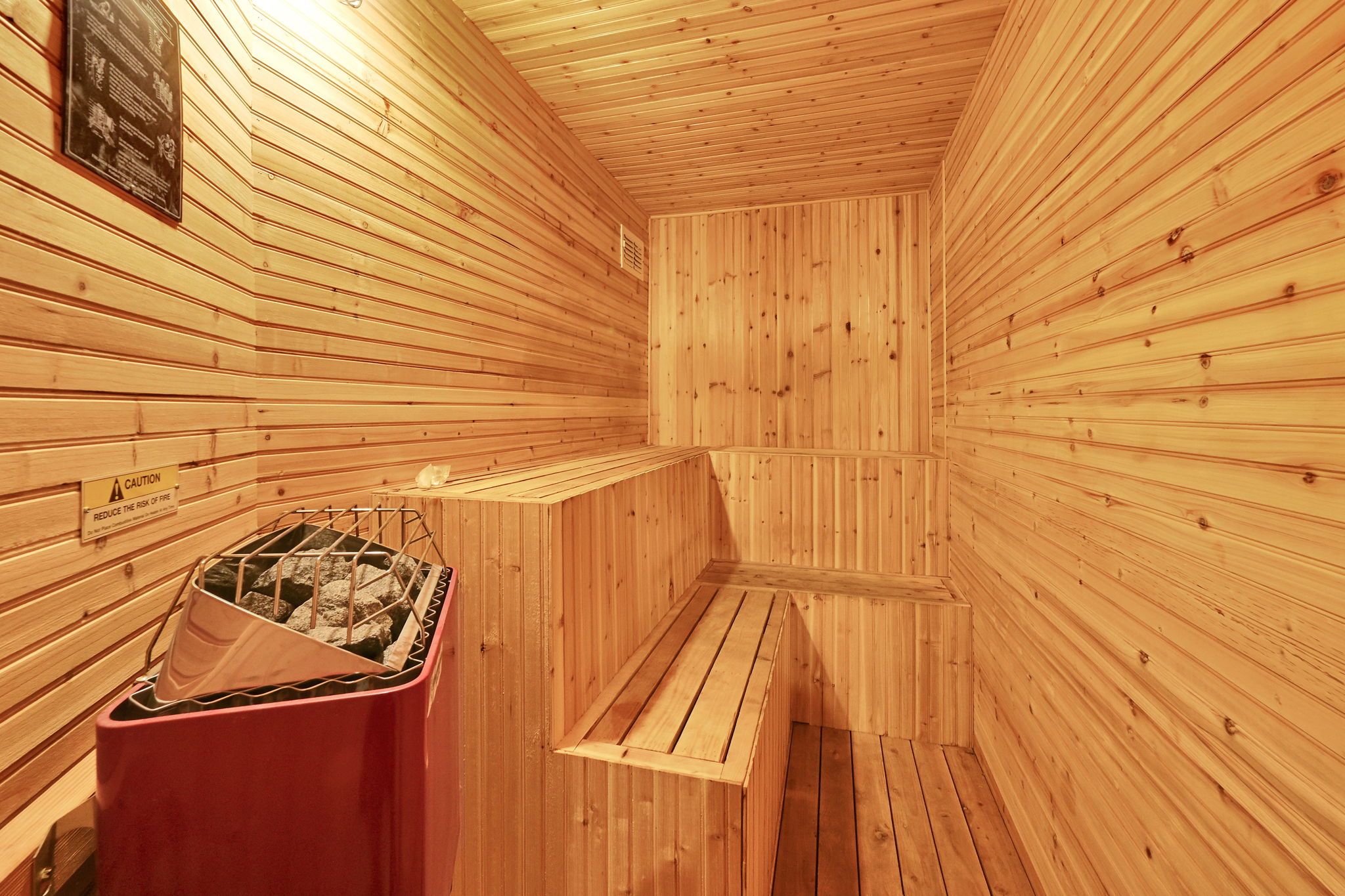 Sauna featuring horizontal wood paneling, multi-level wood benches, a red sauna heater with stones, and overhead lighting