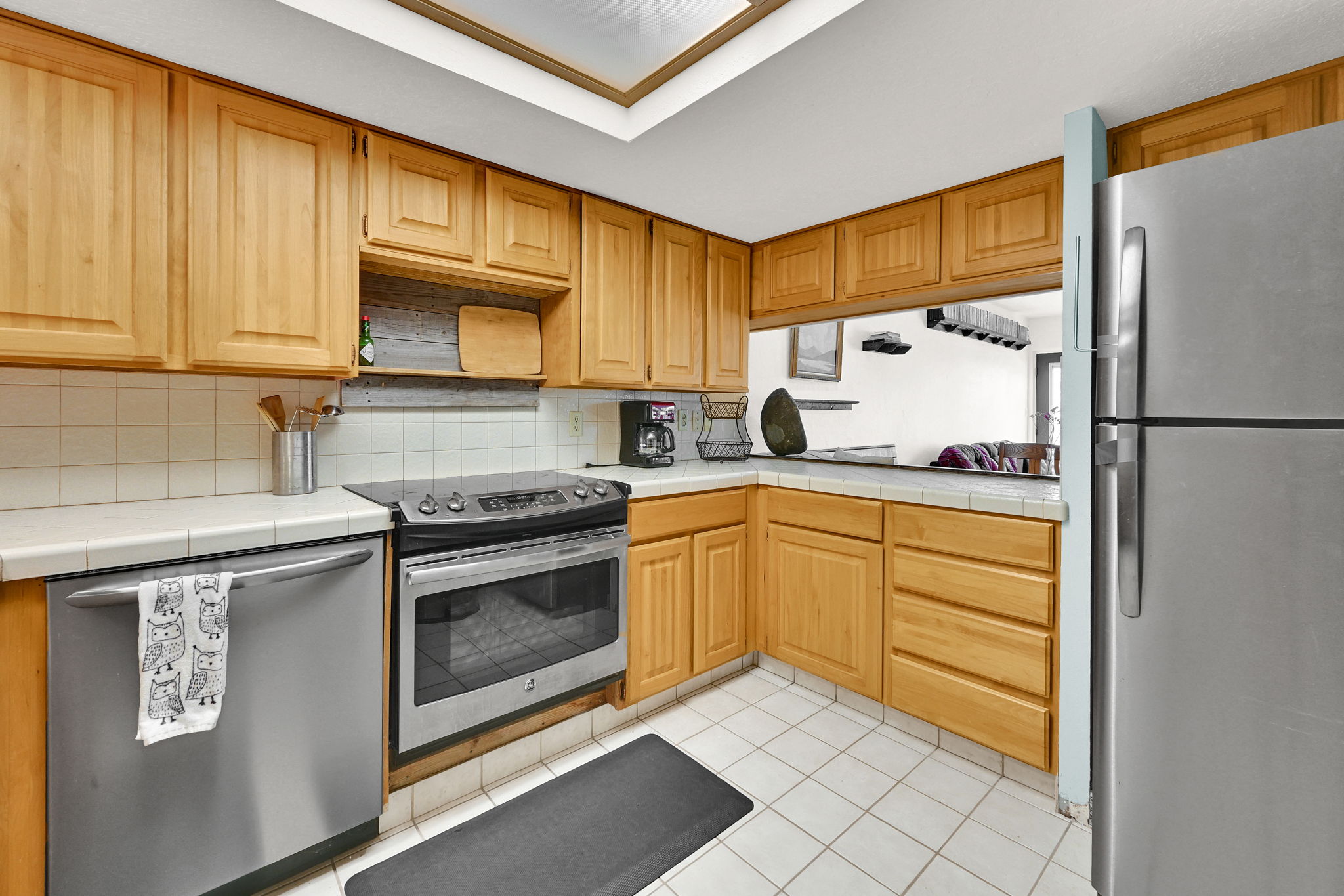 Kitchen featuring wood cabinetry, white tile countertops, stainless steel appliances, white tile flooring, and a skylight