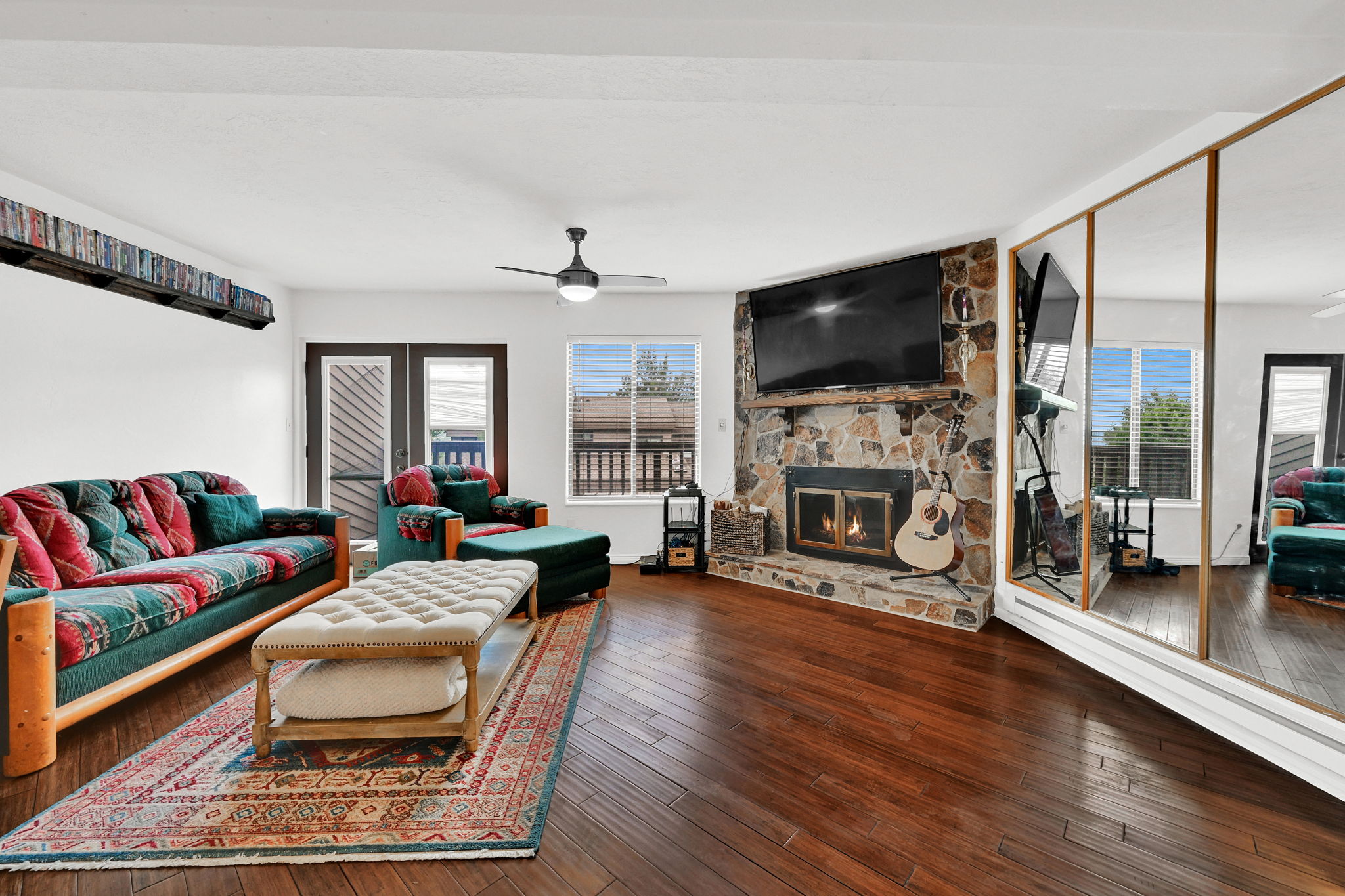 Living room featuring wood-finish flooring, a stone-clad fireplace with hearth, a ceiling fan, and multiple windows