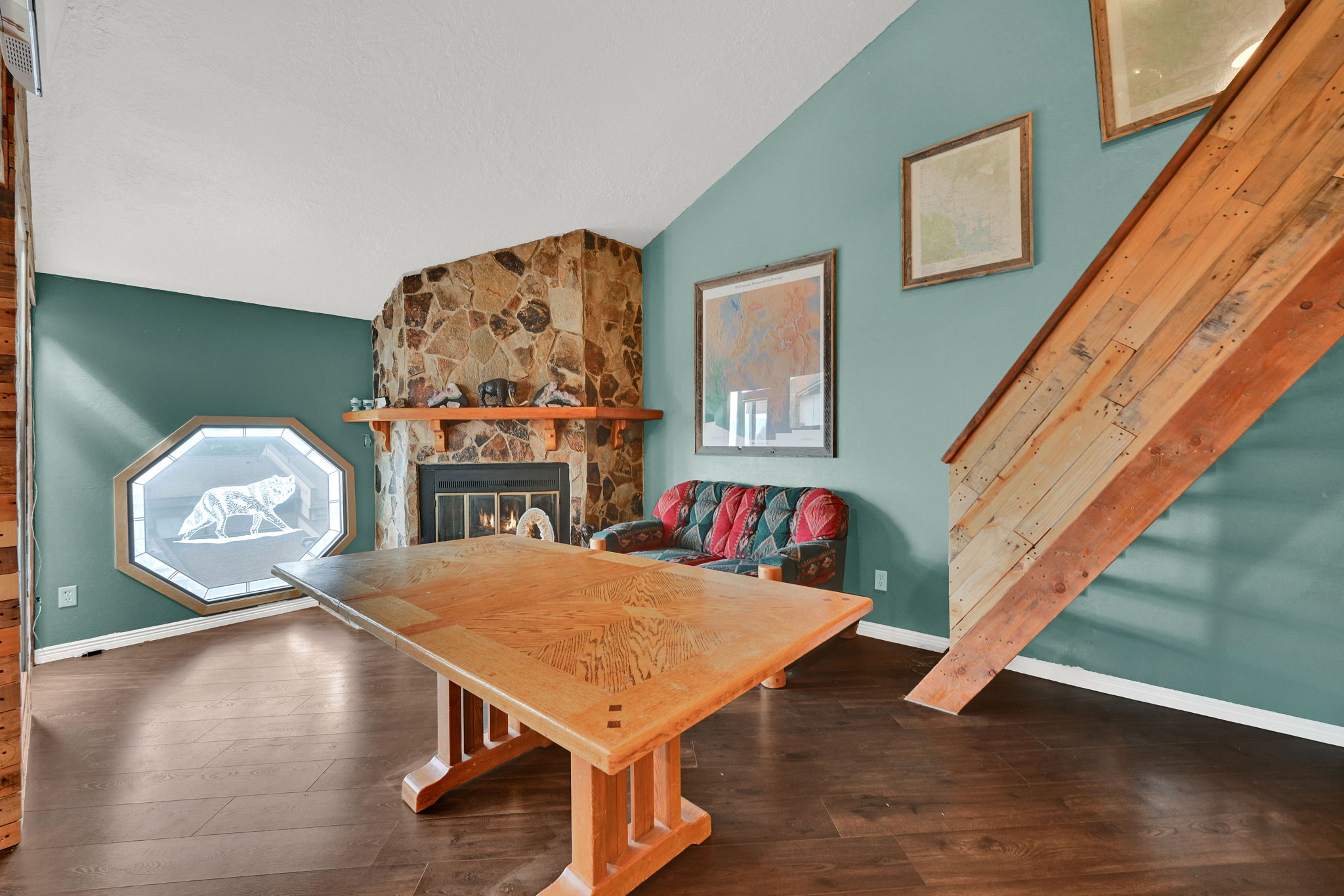 Stone-clad fireplace with mantel, dark wood-finish flooring, vaulted ceiling, octagonal window, and a wood-paneled staircase