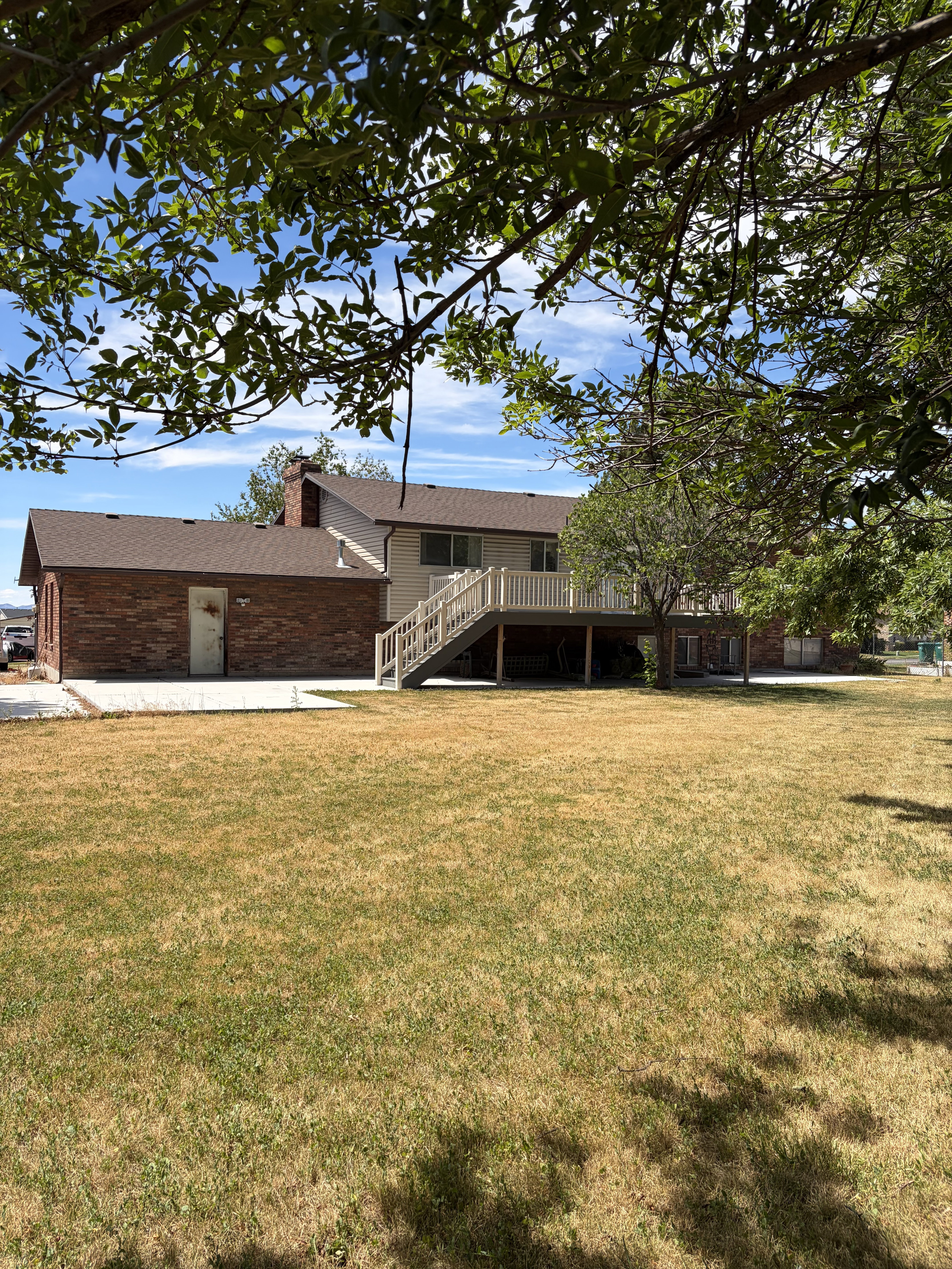 Expansive backyard featuring a large green space, a two-story residence with a brown shingle roof, a brick exterior section, and an elevated deck with a staircase