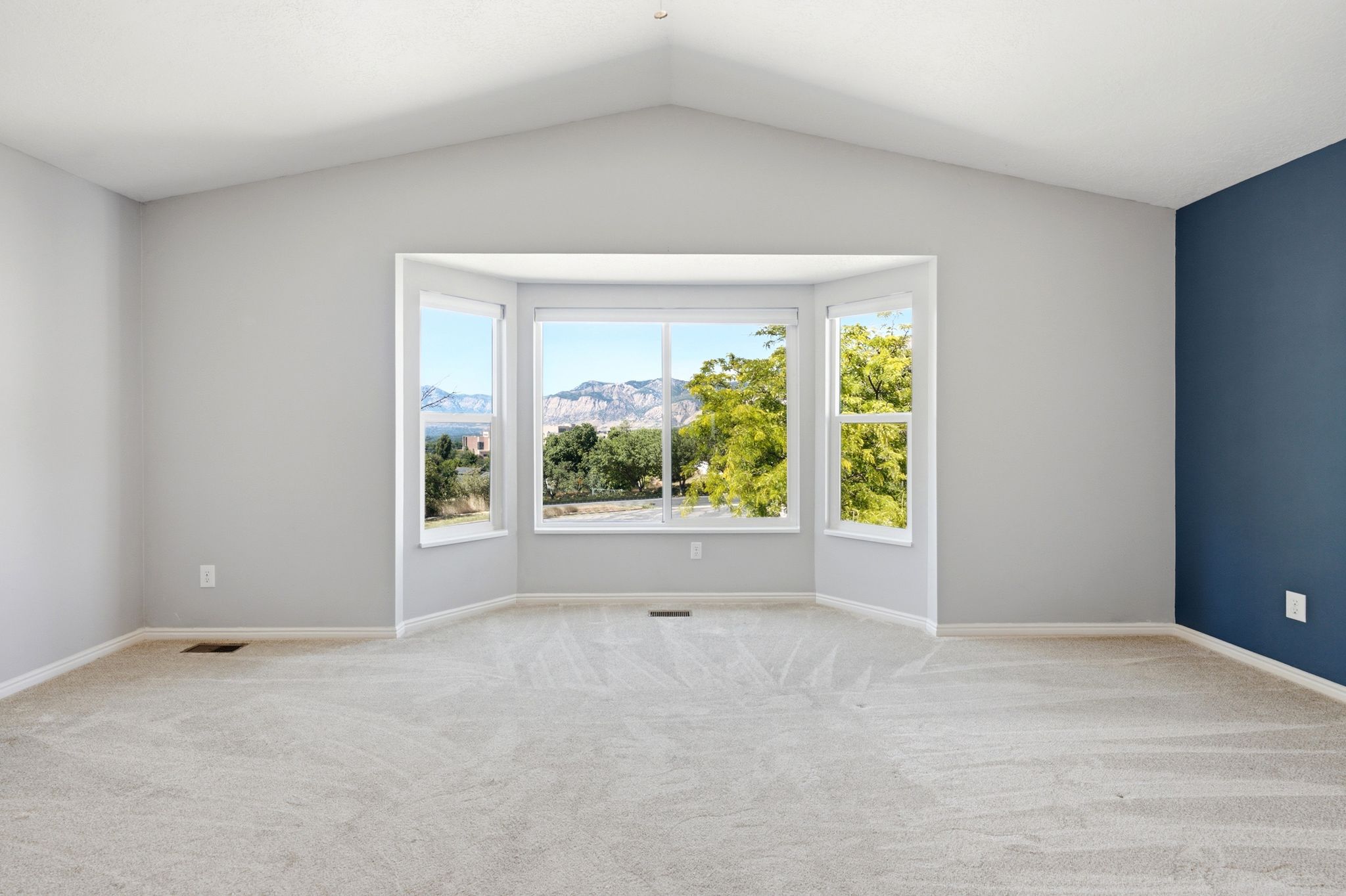 Spacious room featuring vaulted ceilings, bay windows, light-toned carpeting, and a contrasting accent wall, with mountain scenery visible through the windows