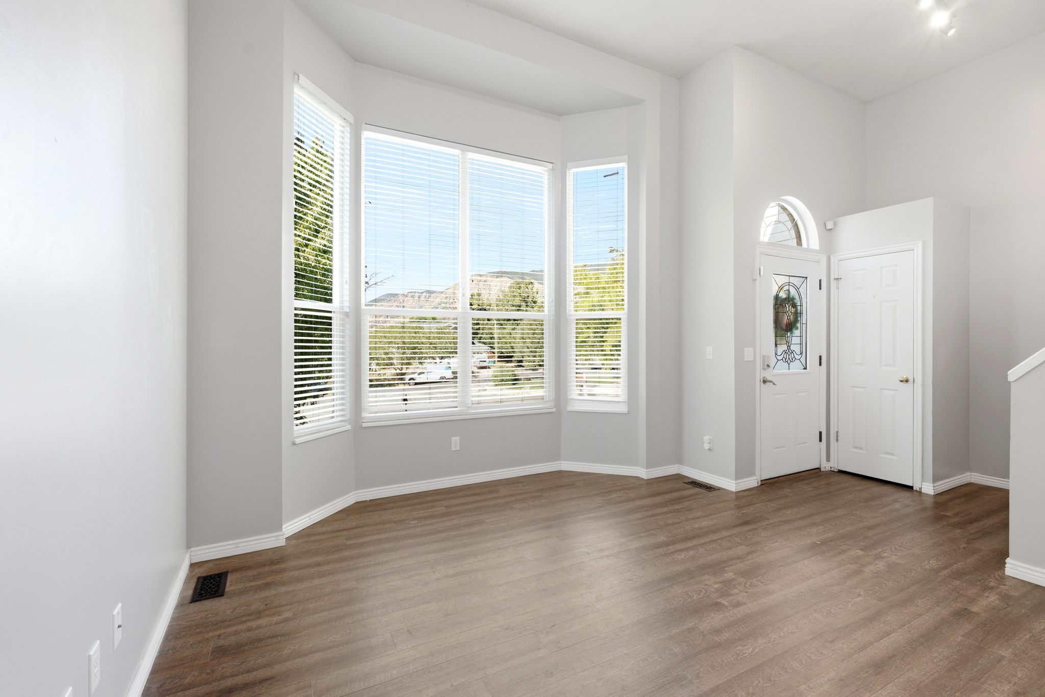 Bay window with multiple large panes, wood-finish flooring, a white front entry door with an arched transom, and light gray wall paint