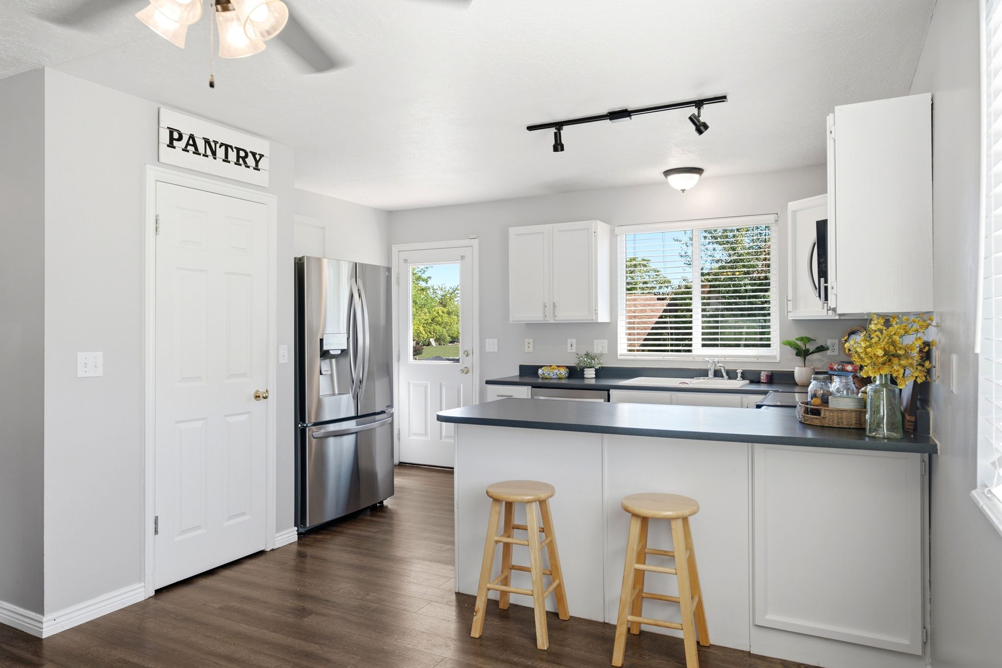 Contemporary kitchen featuring wood-finish flooring, white cabinetry, dark countertops, a stainless steel refrigerator, and a window with horizontal blinds