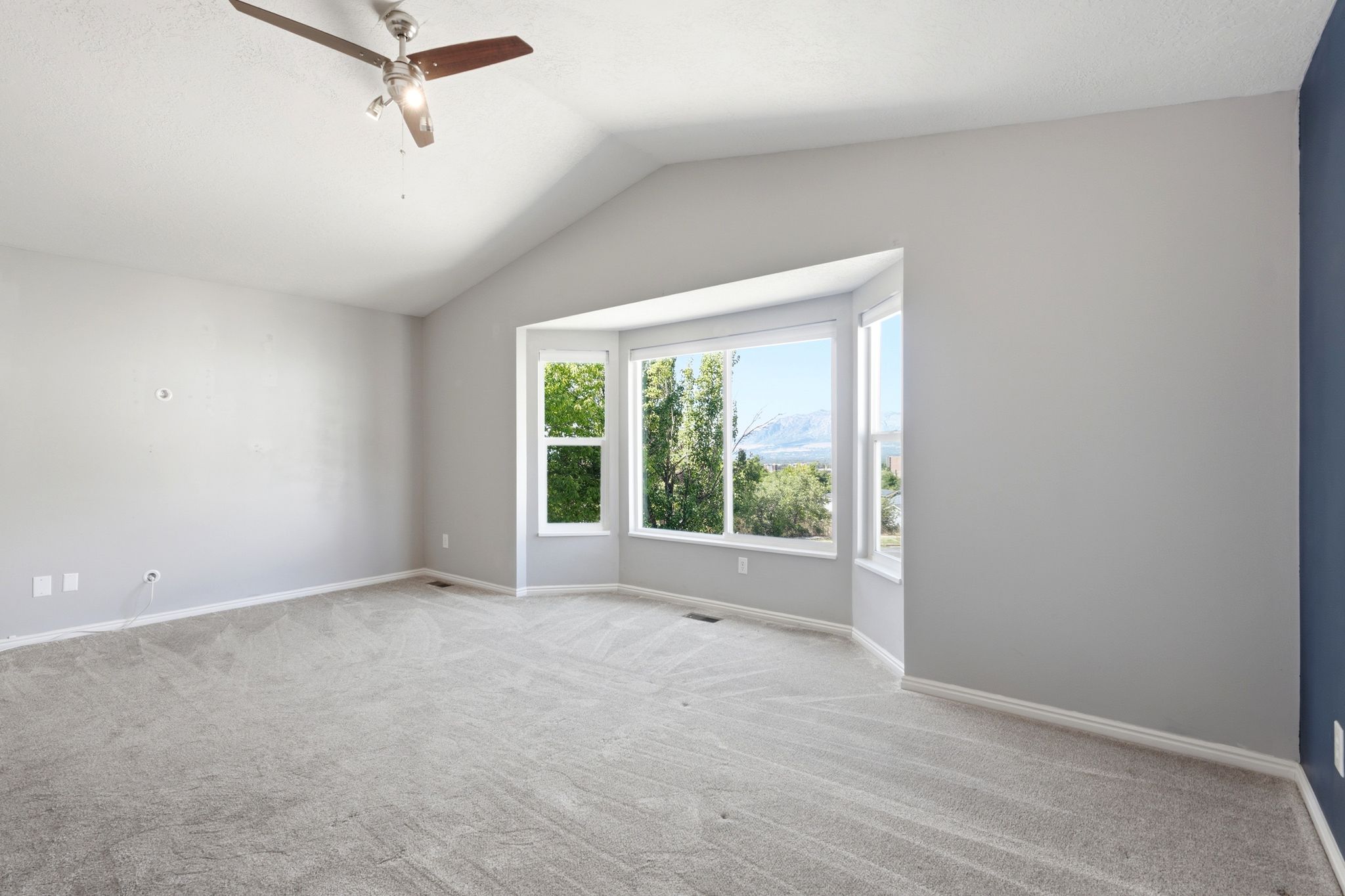 Spacious room featuring vaulted ceilings, a prominent bay window, light gray wall paint, and a ceiling fan with integrated lighting