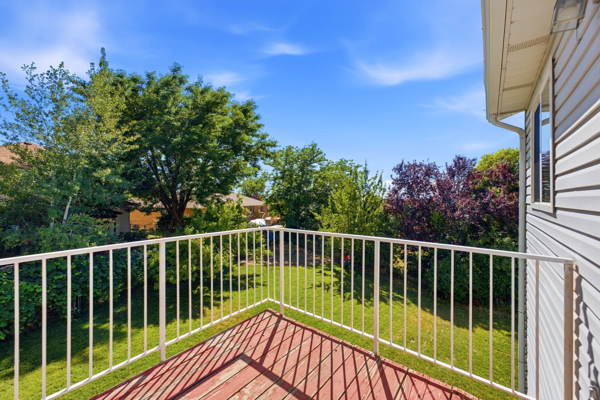 Spacious outdoor deck with wood-finish flooring and a white railing, overlooking a verdant lawn and mature landscaping