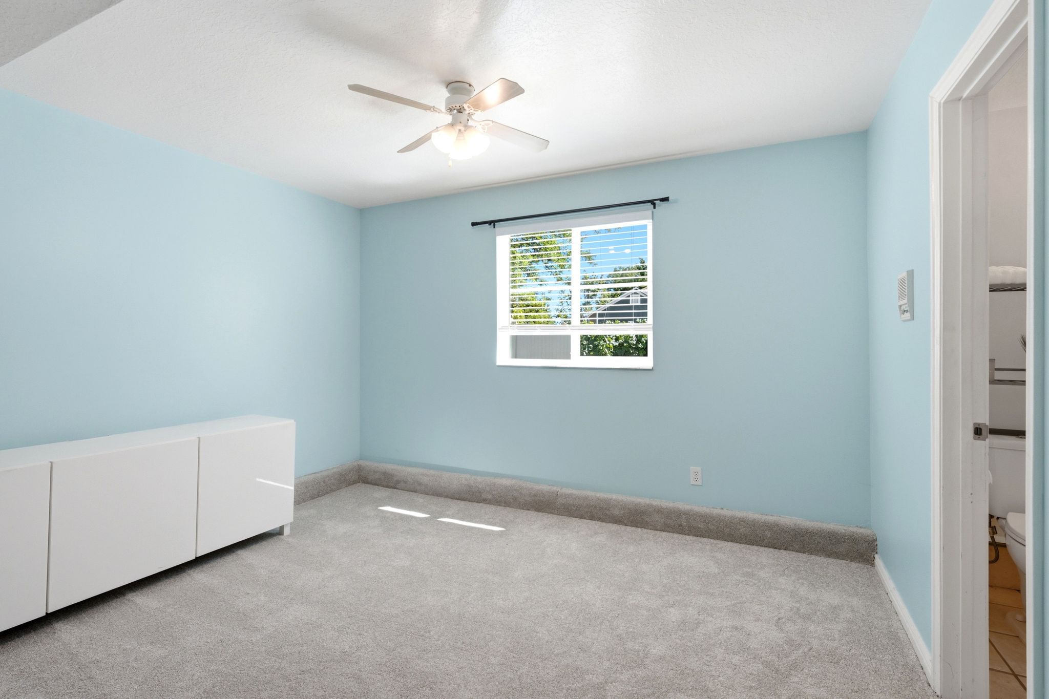 Bright room featuring a window with blinds, a ceiling fan, light blue walls, and grey carpeting