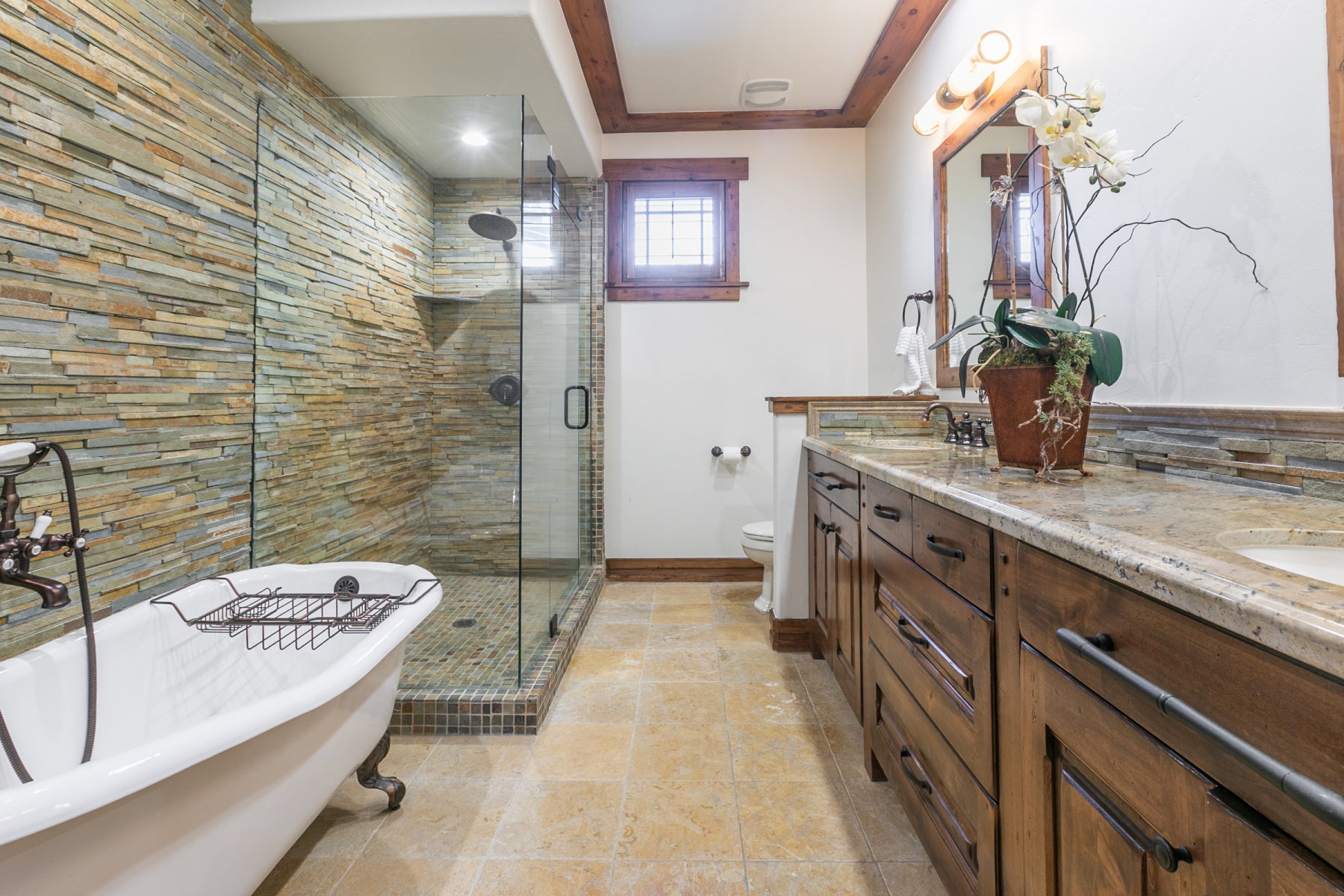 Bathroom with a stone accent wall, glass-enclosed shower, freestanding clawfoot tub, dual vanity with granite countertops, and wood-finish cabinetry