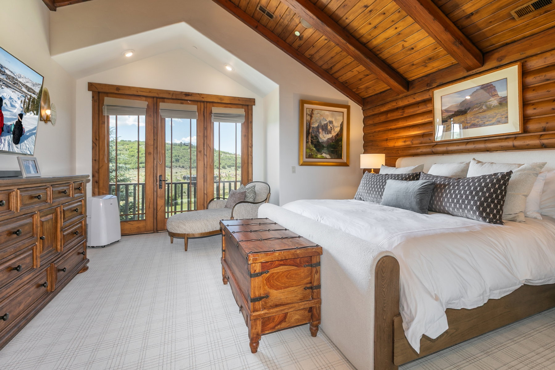 Spacious room featuring a vaulted wood-beam ceiling, light-toned patterned carpet, a full-length triple-panel door with a private balcony, and a log-style accent wall