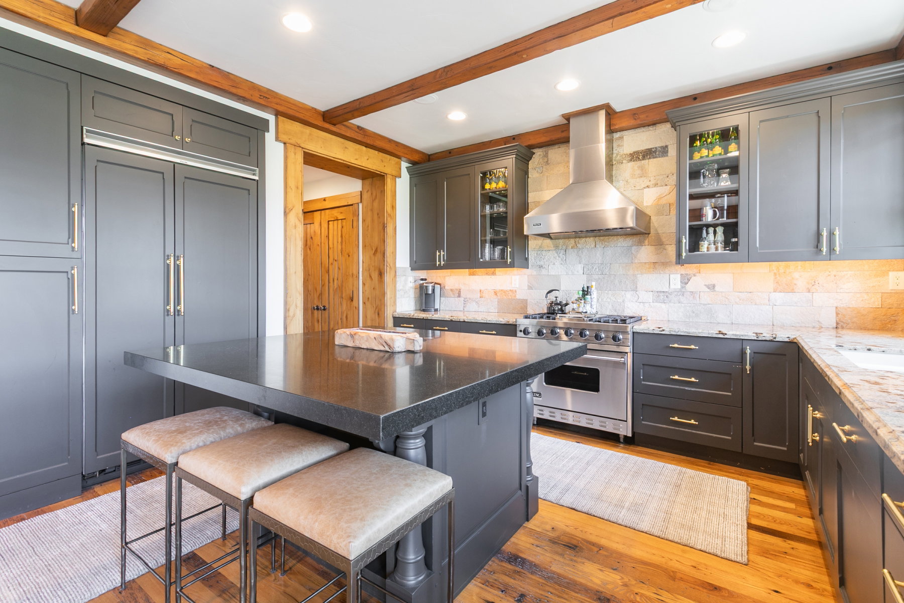Gourmet kitchen featuring dark gray cabinetry, exposed wood ceiling beams, wood-finish flooring, a stainless steel range with matching hood, and stone countertops with a coordinating tile backsplash