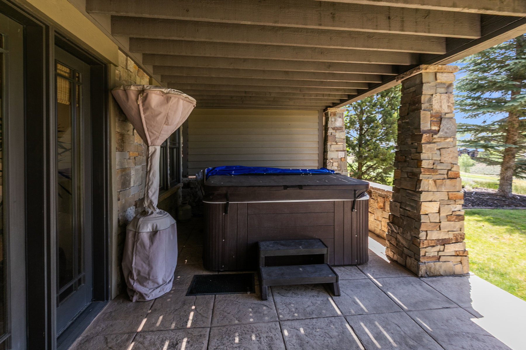Covered patio with stamped concrete flooring, featuring a built-in spa with a protective cover, stone veneer columns, and natural wood ceiling beams