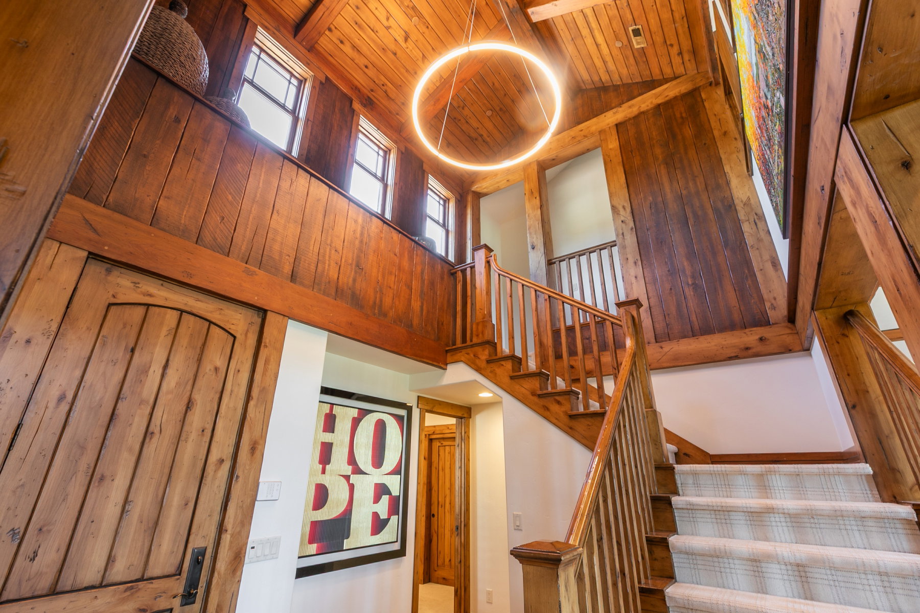 Vaulted wood-paneled ceiling with exposed beams, wood-finish flooring staircase with carpet runner, circular modern chandelier, and natural wood doors
