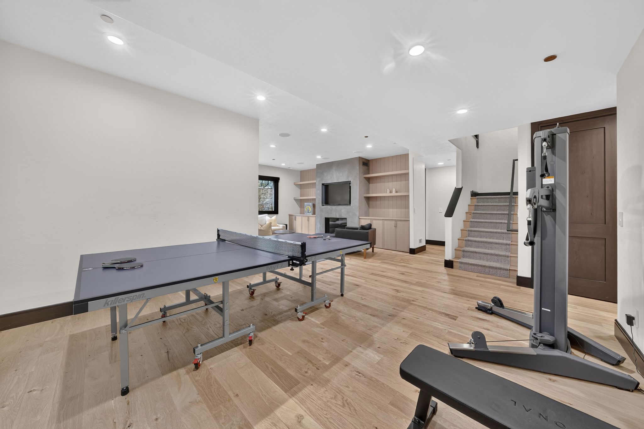 Expansive recreation room featuring light wood-finish flooring, recessed lighting, a built-in media center with shelving, and a carpeted staircase with a dark wood railing