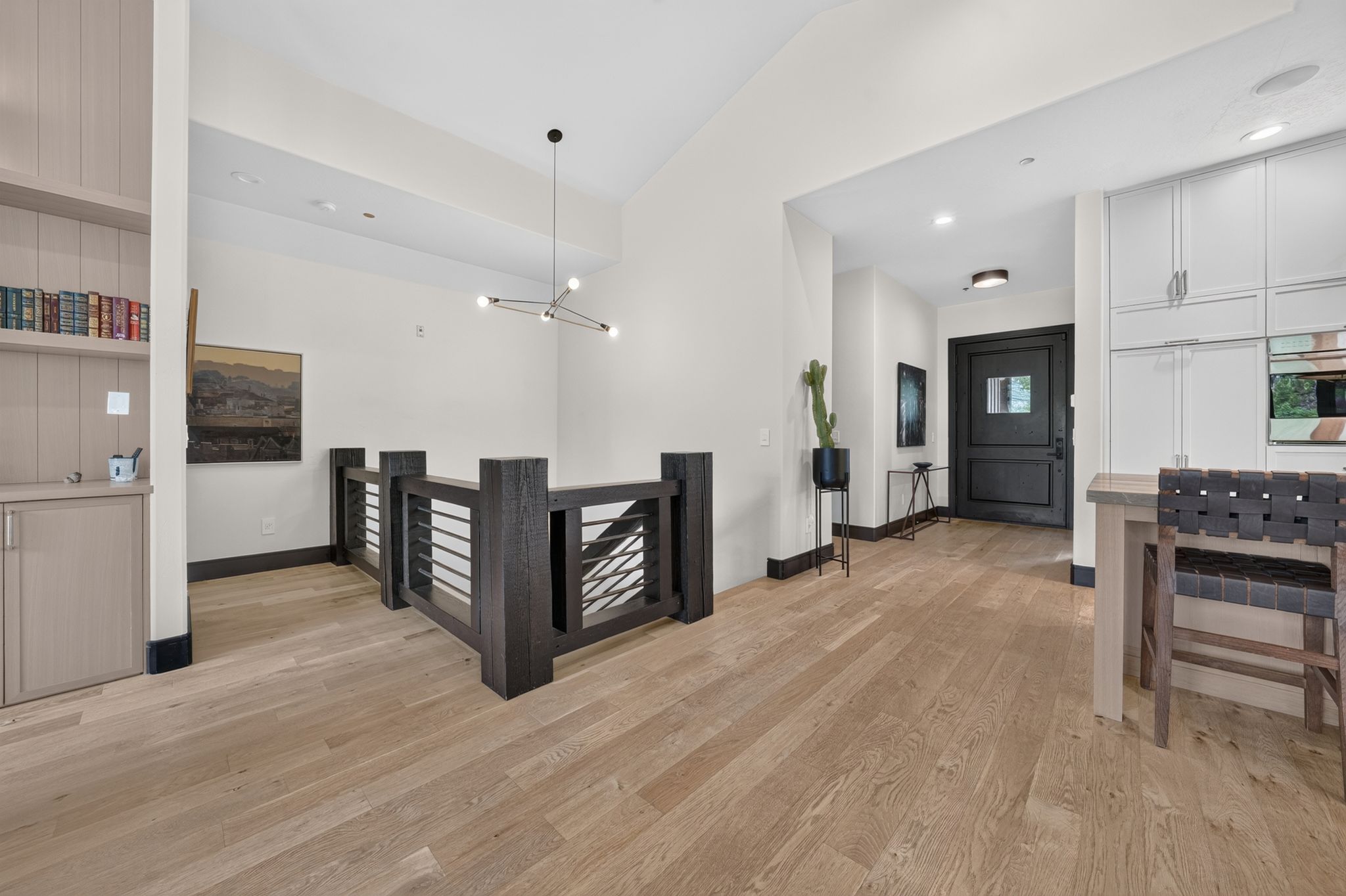 Open-concept living area with light wood-finish flooring, a dark wood and metal railing, a built-in shelving unit with lower cabinetry, a built-in kitchen area with white cabinetry, and a dark entry door