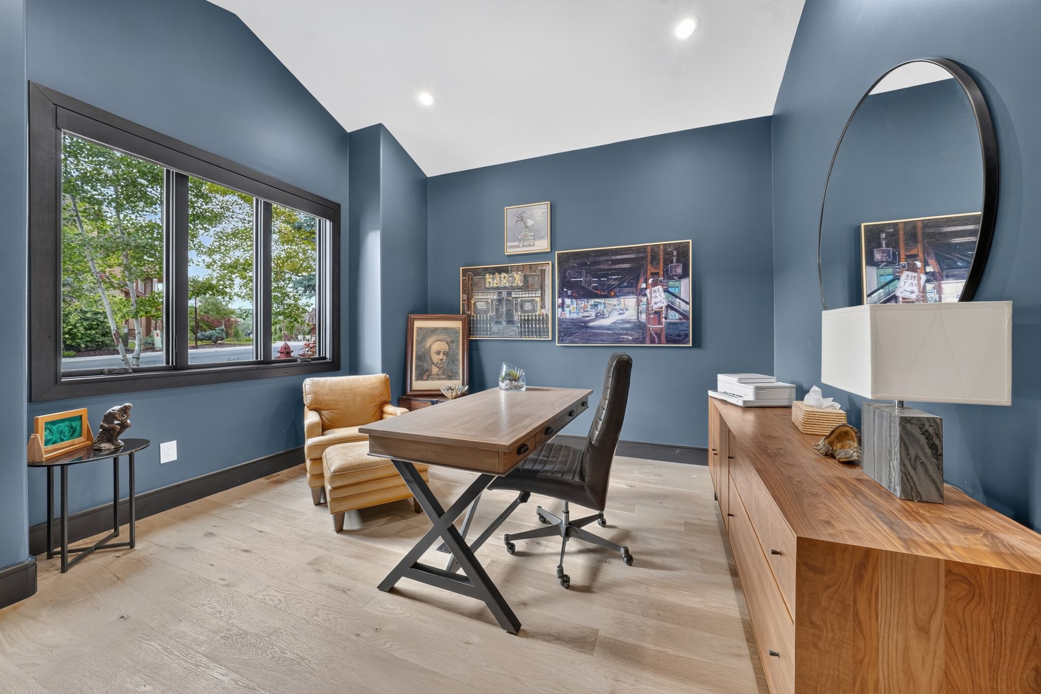 Spacious room featuring light wood-finish flooring, dark trim accents, recessed lighting, and a large window overlooking mature trees