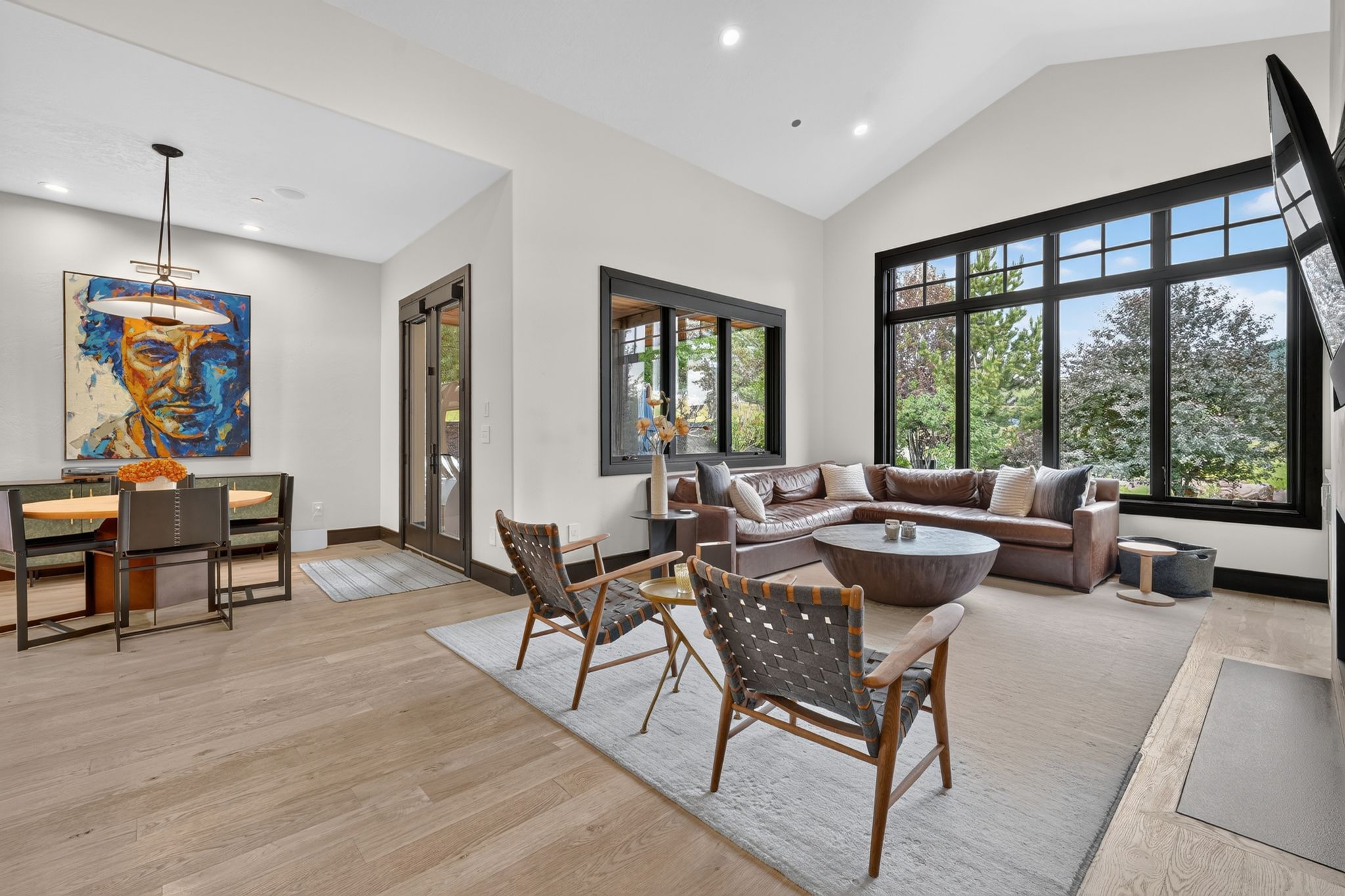 Vaulted ceilings frame the open concept living area featuring large windows with dark trim, light wood-finish flooring, and recessed lighting