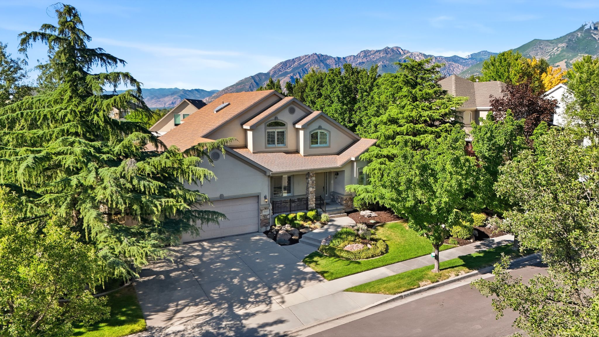 Traditional stucco and stone exterior featuring a two-car garage, mature trees, and a landscaped front yard with a paved driveway