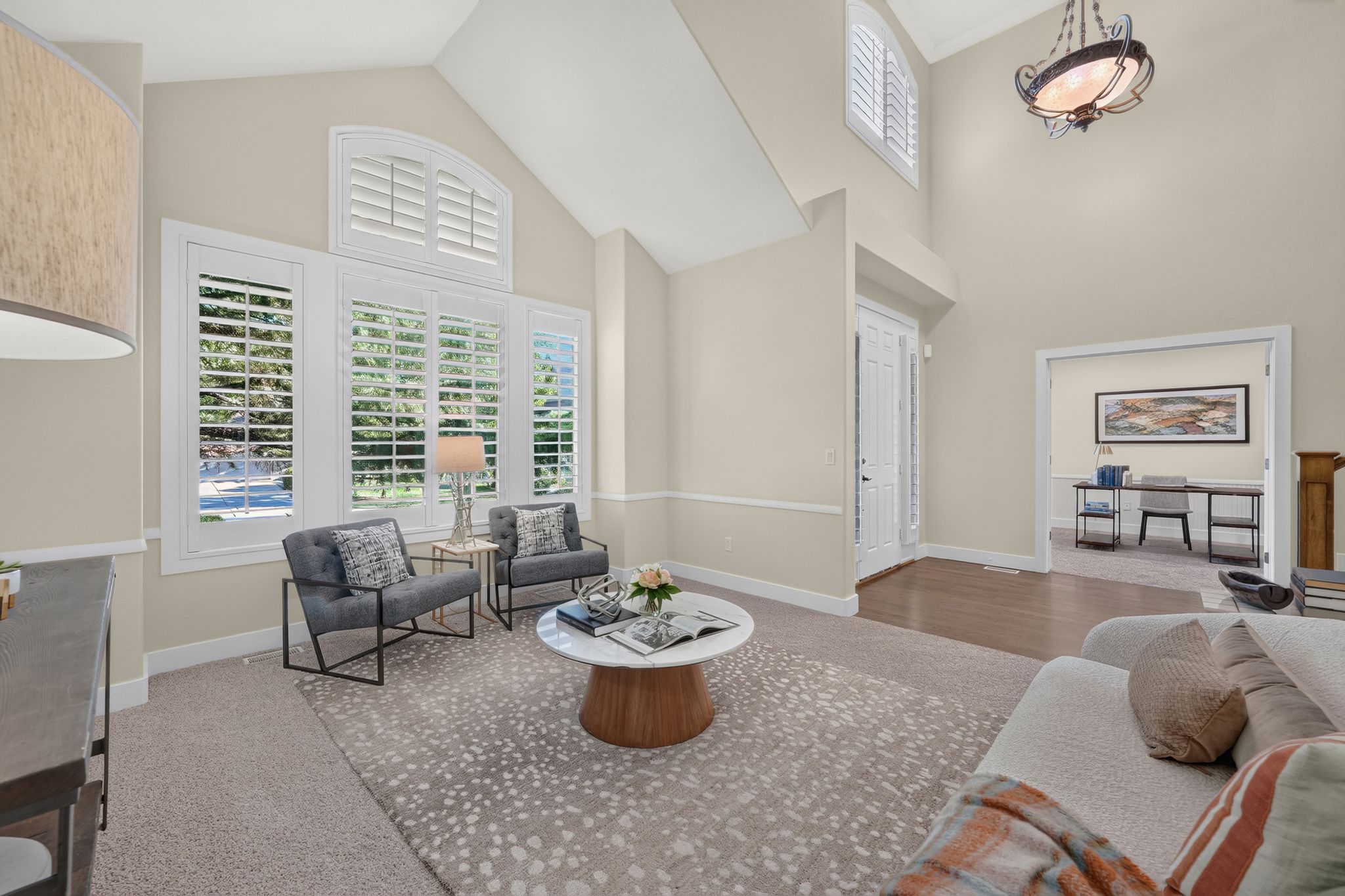 Vaulted ceiling living area featuring a prominent window wall with plantation shutters, a double-door entryway, and hardwood flooring accents