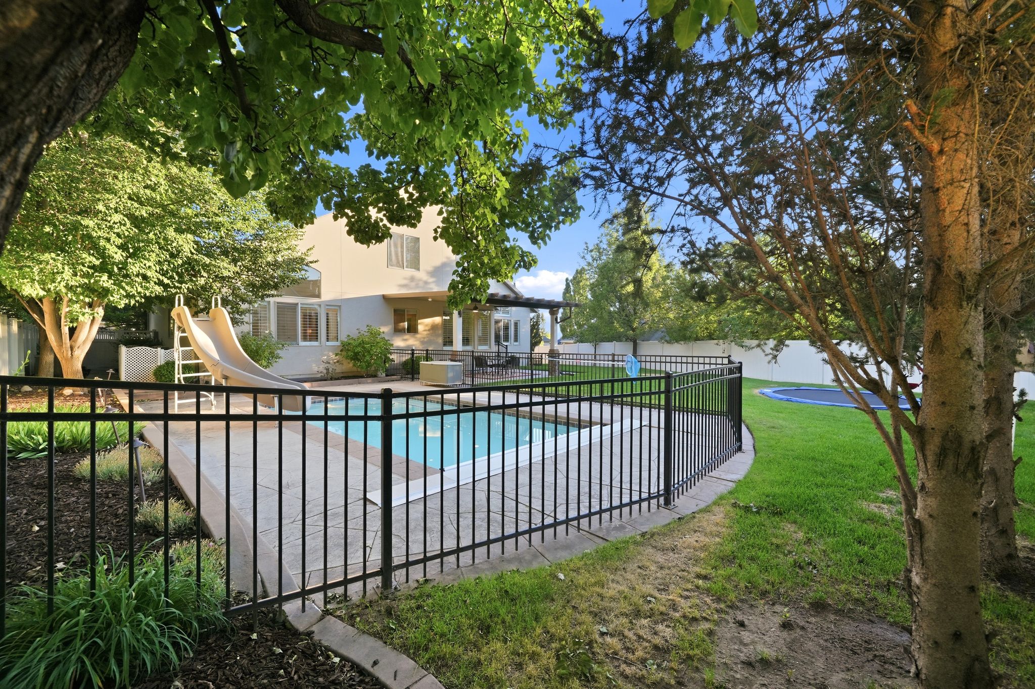 Outdoor swimming pool with surrounding concrete patio, black wrought iron-style fencing, and a grassy yard