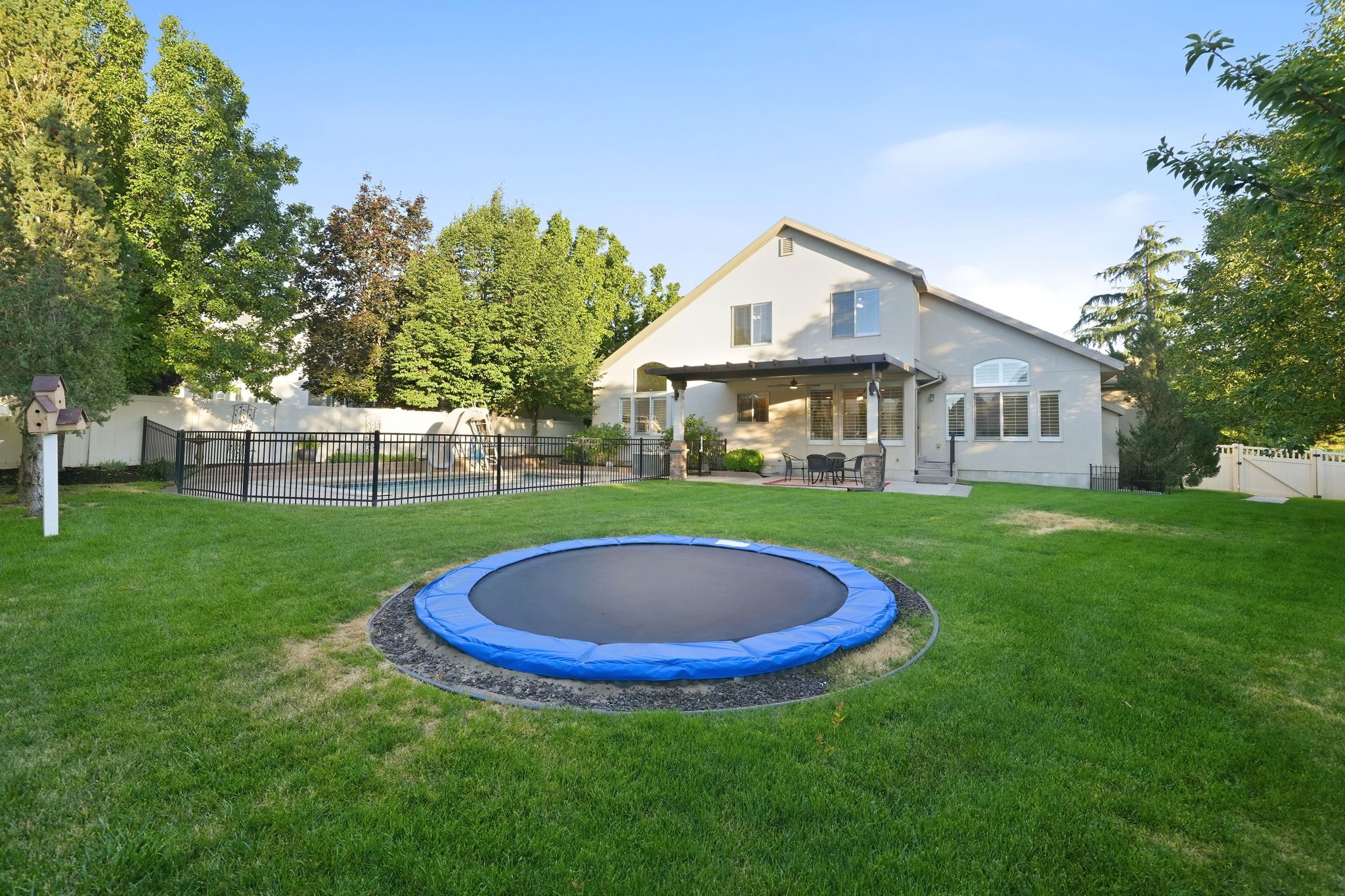 Expansive backyard featuring a manicured lawn, a fenced swimming pool, mature perimeter trees, and a covered patio area with outdoor seating