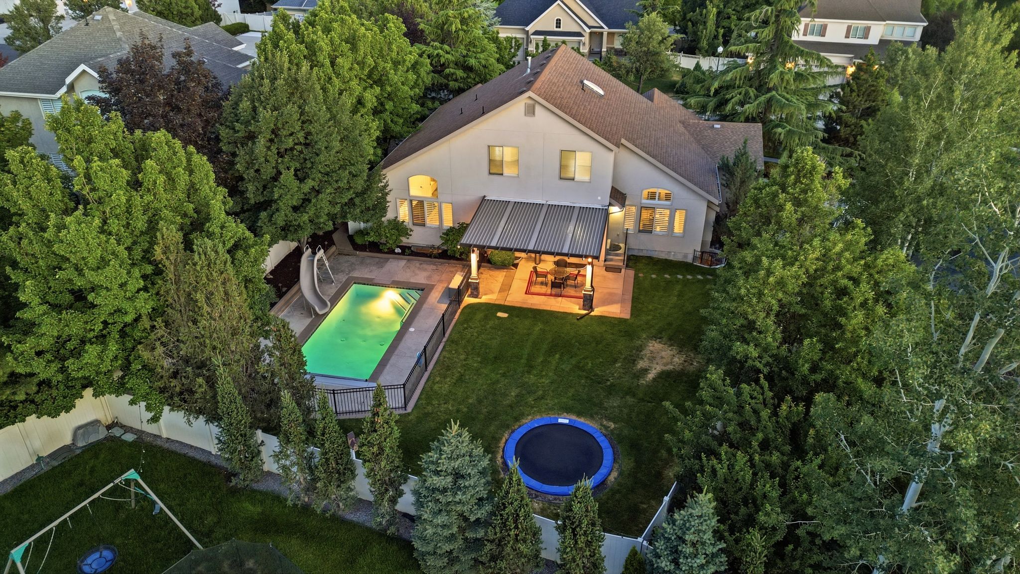 Rear exterior featuring a rectangular in-ground swimming pool with perimeter fencing and an attached slide, a covered patio area with recessed lighting, a manicured lawn, mature trees, and a privacy fence