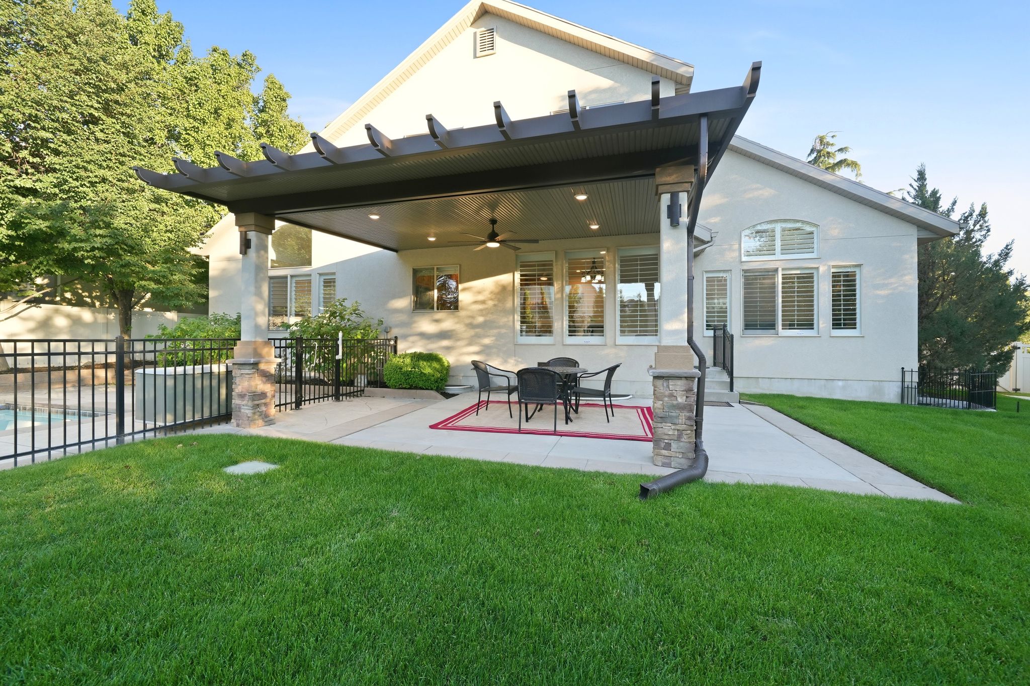 Covered patio with a built-in ceiling fan, recessed lighting, and stone-clad pillars, overlooking a fenced swimming pool and a manicured lawn