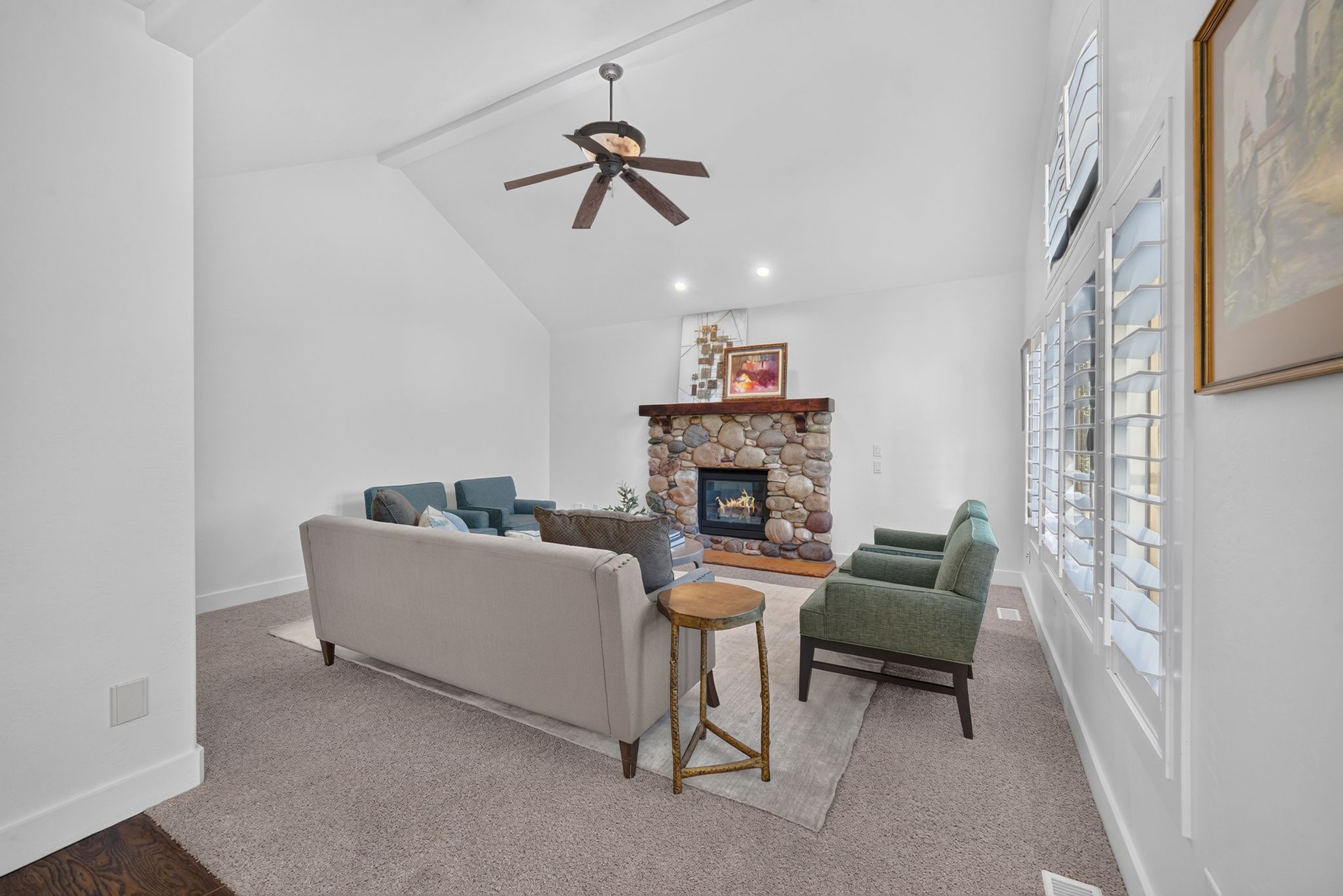 Living area featuring a floor-to-ceiling stone fireplace with a wood mantle, vaulted ceiling with a ceiling fan, and extensive windows with plantation shutters