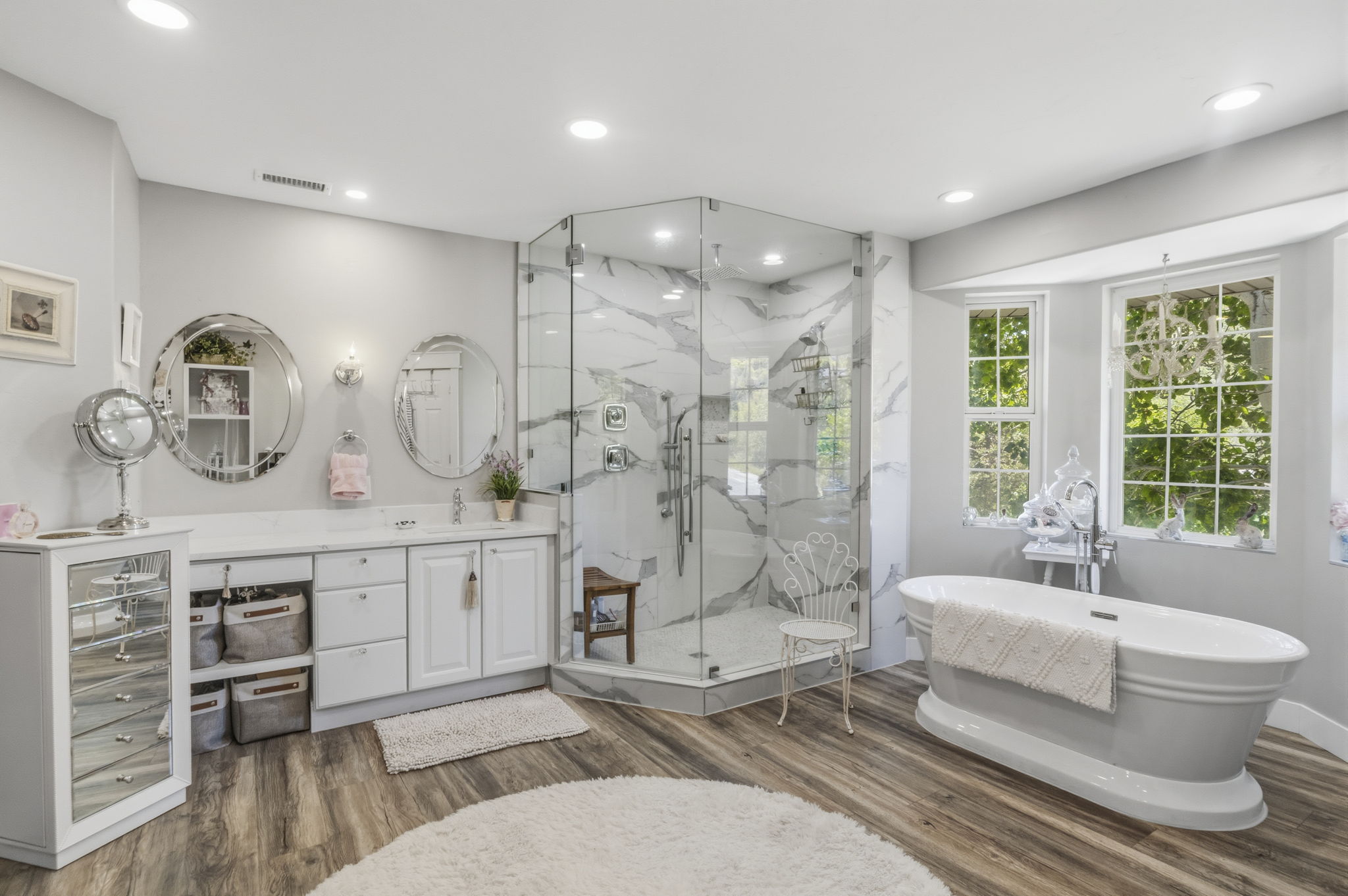 Spacious bath featuring a freestanding tub, expansive glass-enclosed shower with marble-look tile, double vanity with white cabinetry and countertop, and wood-finish flooring