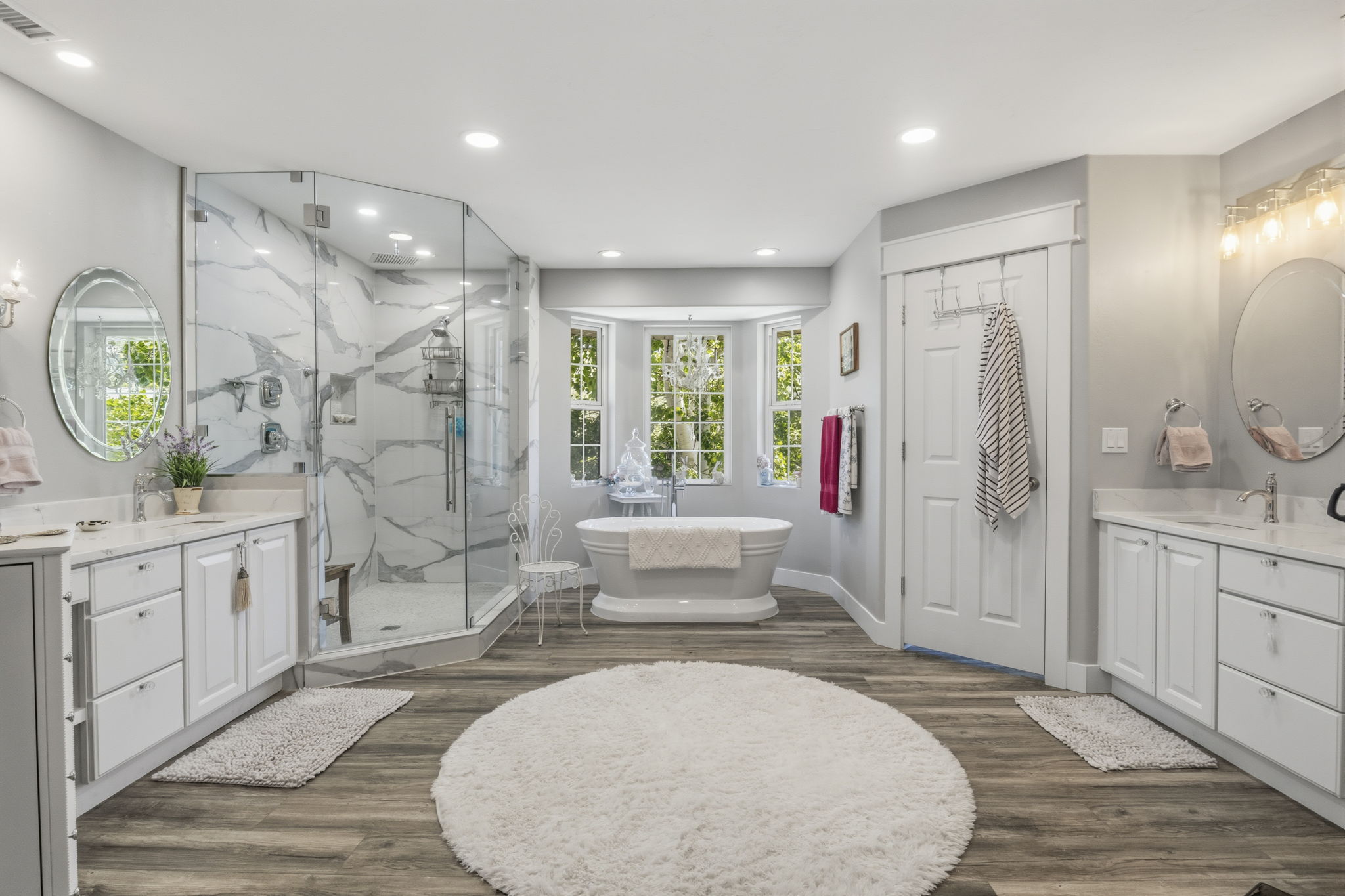 Spacious bathroom featuring wood-finish flooring, a freestanding soaking tub in a bay window, and a glass-enclosed shower with marble-look wall tiling