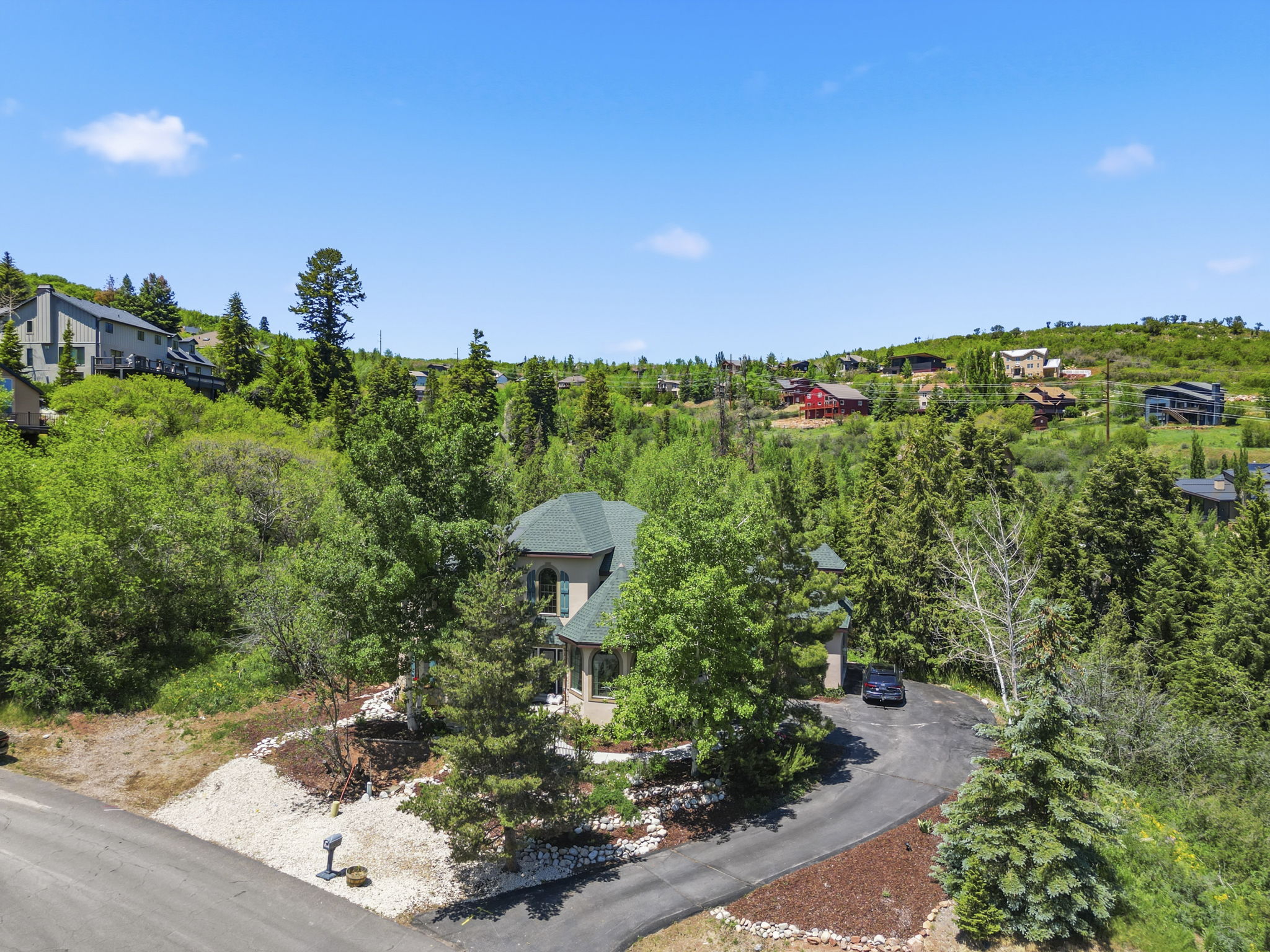 Elevated property surrounded by mature trees, featuring a paved driveway, and a natural landscape design with decorative rock and mulch