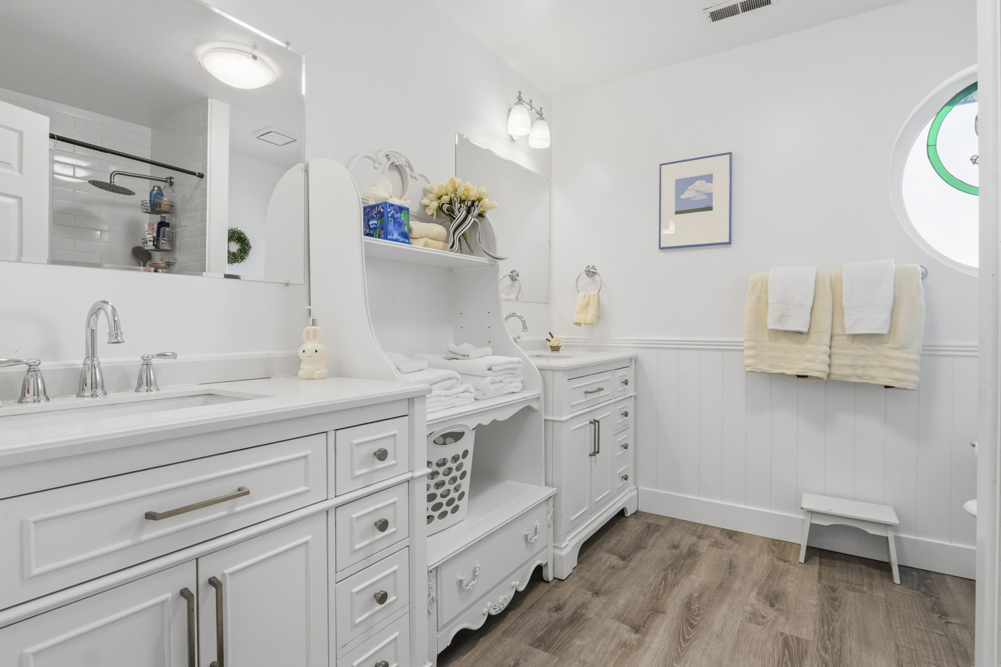 Bright full bathroom featuring a dual vanity with white cabinetry, wood-finish flooring, a glass-enclosed shower, and a circular window