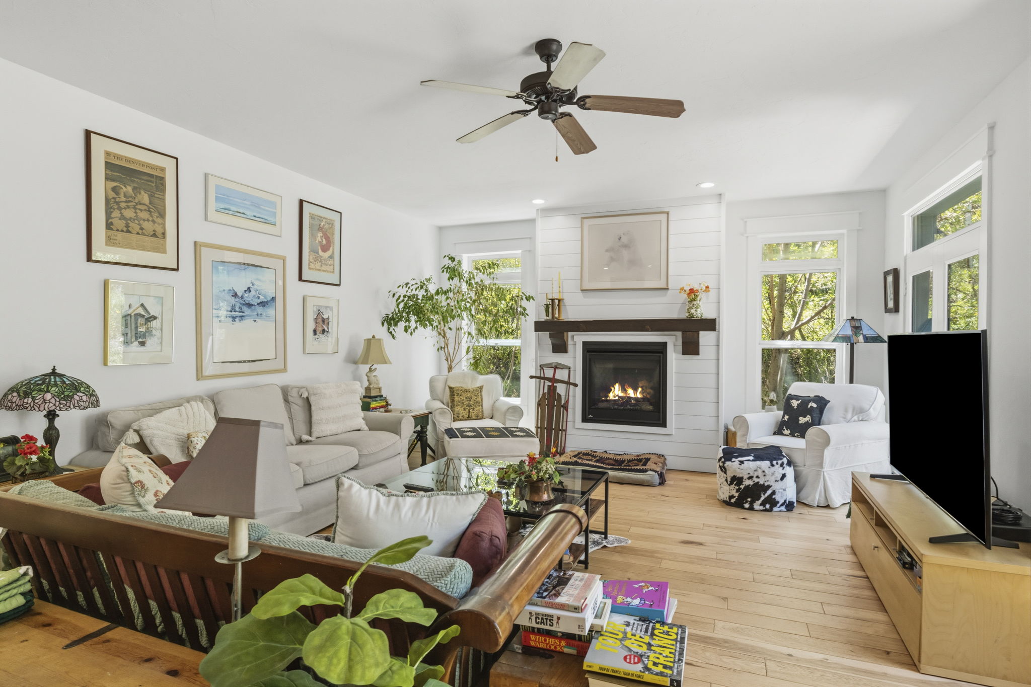 Spacious living area featuring a central fireplace with wood mantel, shiplap accent wall, wood-finish flooring, ceiling fan, and multiple windows