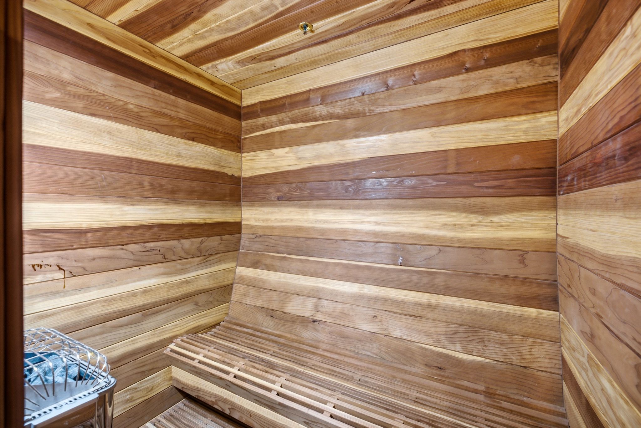 Wood-paneled sauna featuring a built-in slatted bench, stainless steel heater, and ceiling-mounted sprinkler head