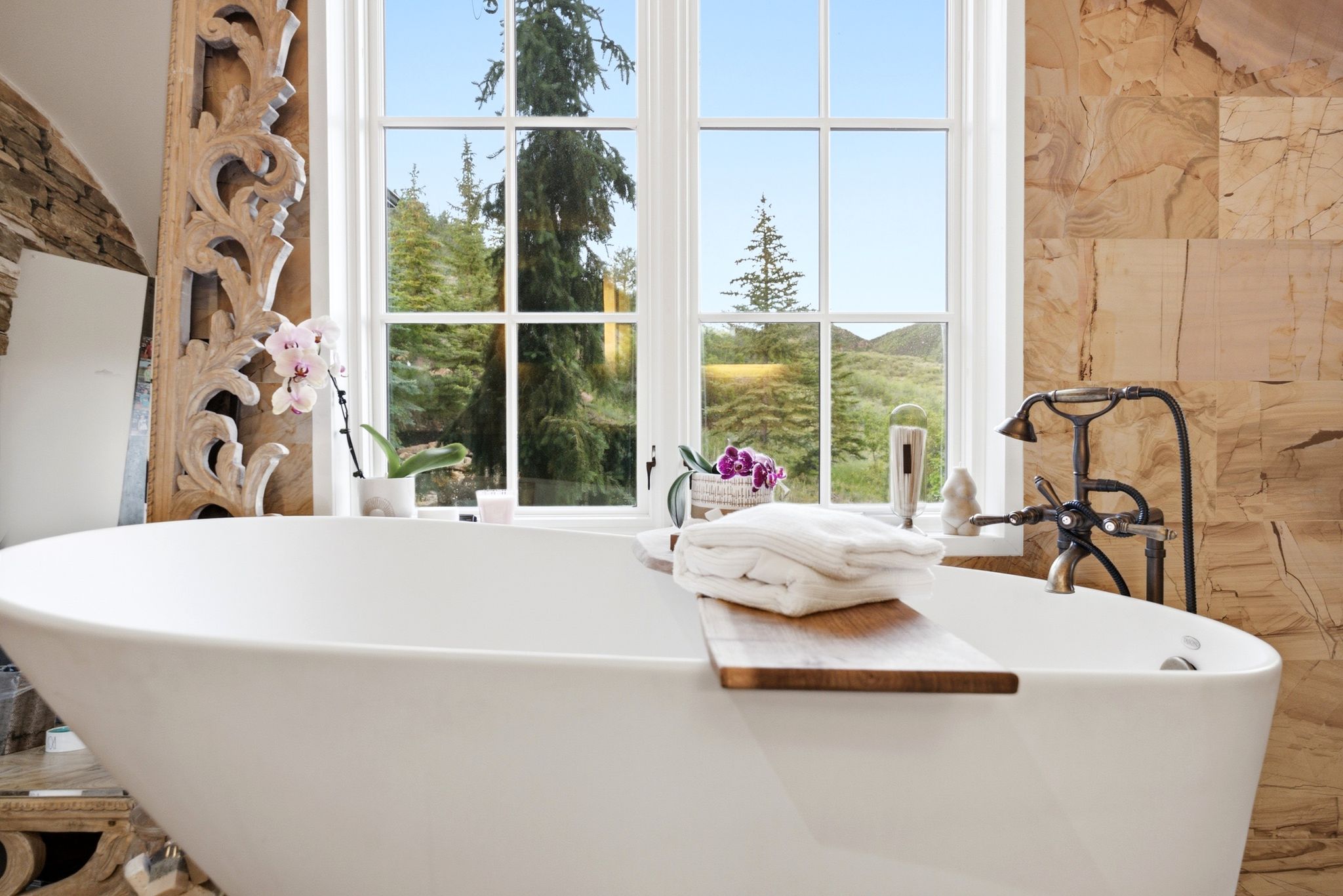 Freestanding soaking tub with an antique-finish tub filler, white window framing natural scenery, and a stone-tiled wall