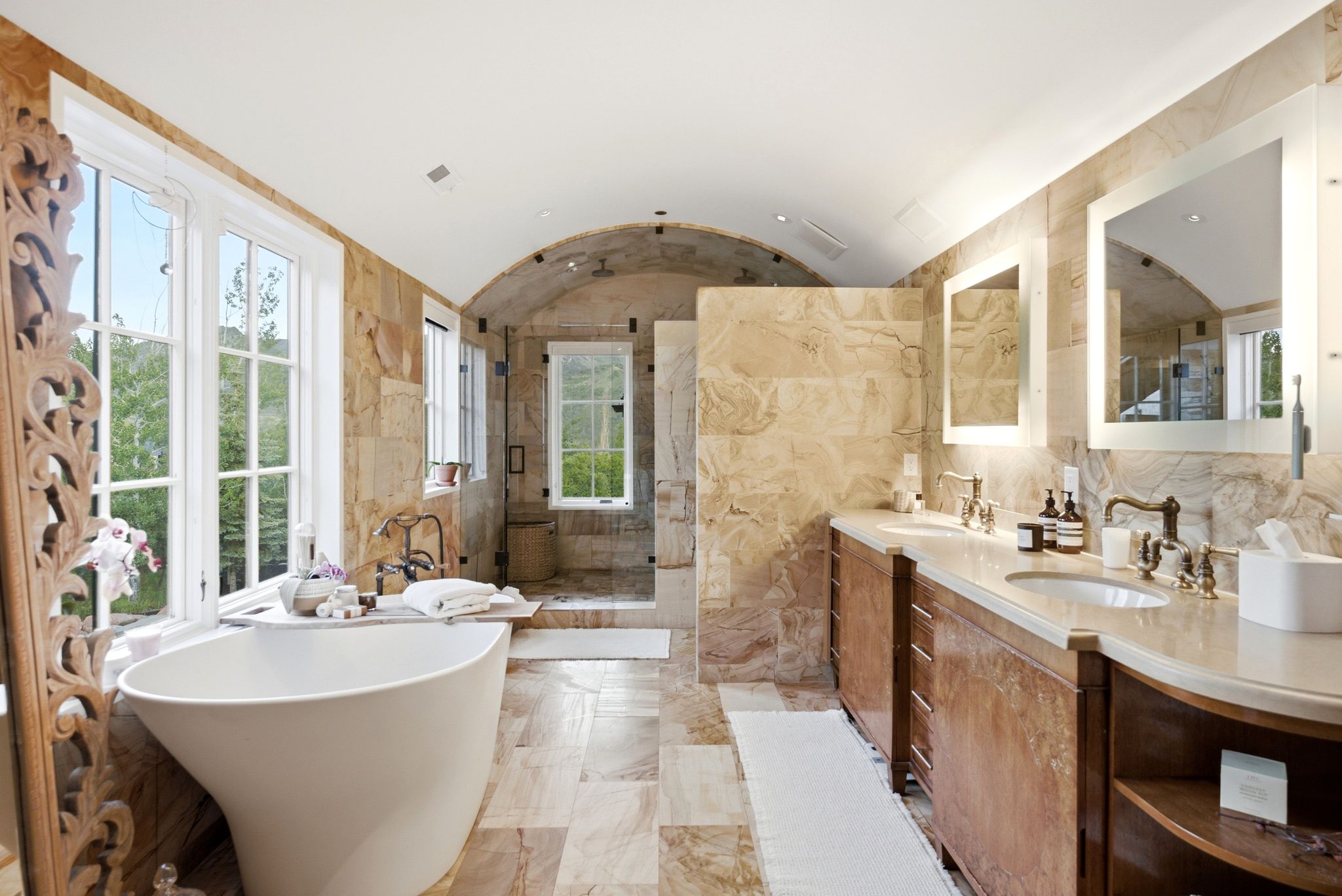 Spacious bathroom featuring a freestanding soaking tub, dual vanity with undermount sinks, stone tile flooring, arched ceiling, and large windows