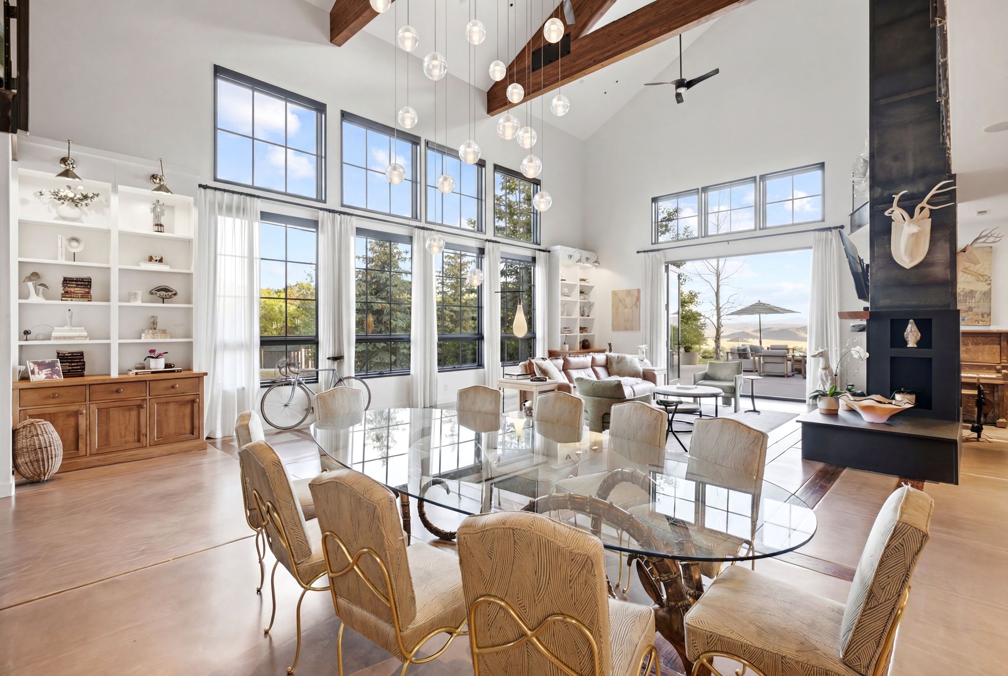 Vaulted ceilings with exposed timber beams, expansive black-framed windows, built-in shelving, glass dining table, and a contemporary fireplace feature