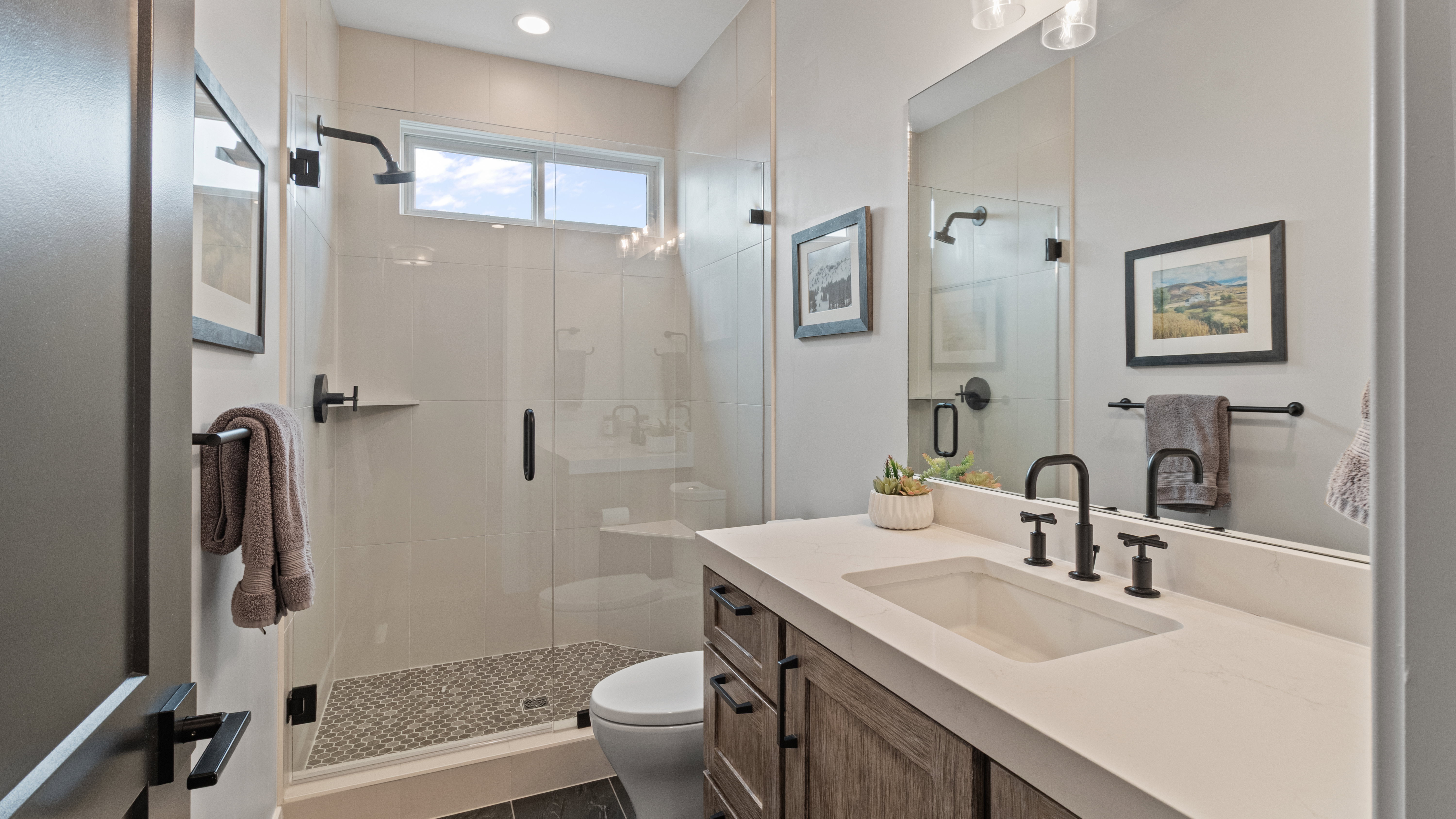 Main level 2nd bathroom featuring a spacious glass-enclosed shower with mosaic tile flooring, a custom wood-finish vanity with an integrated sink, matte black fixtures, and a transom window for natural light