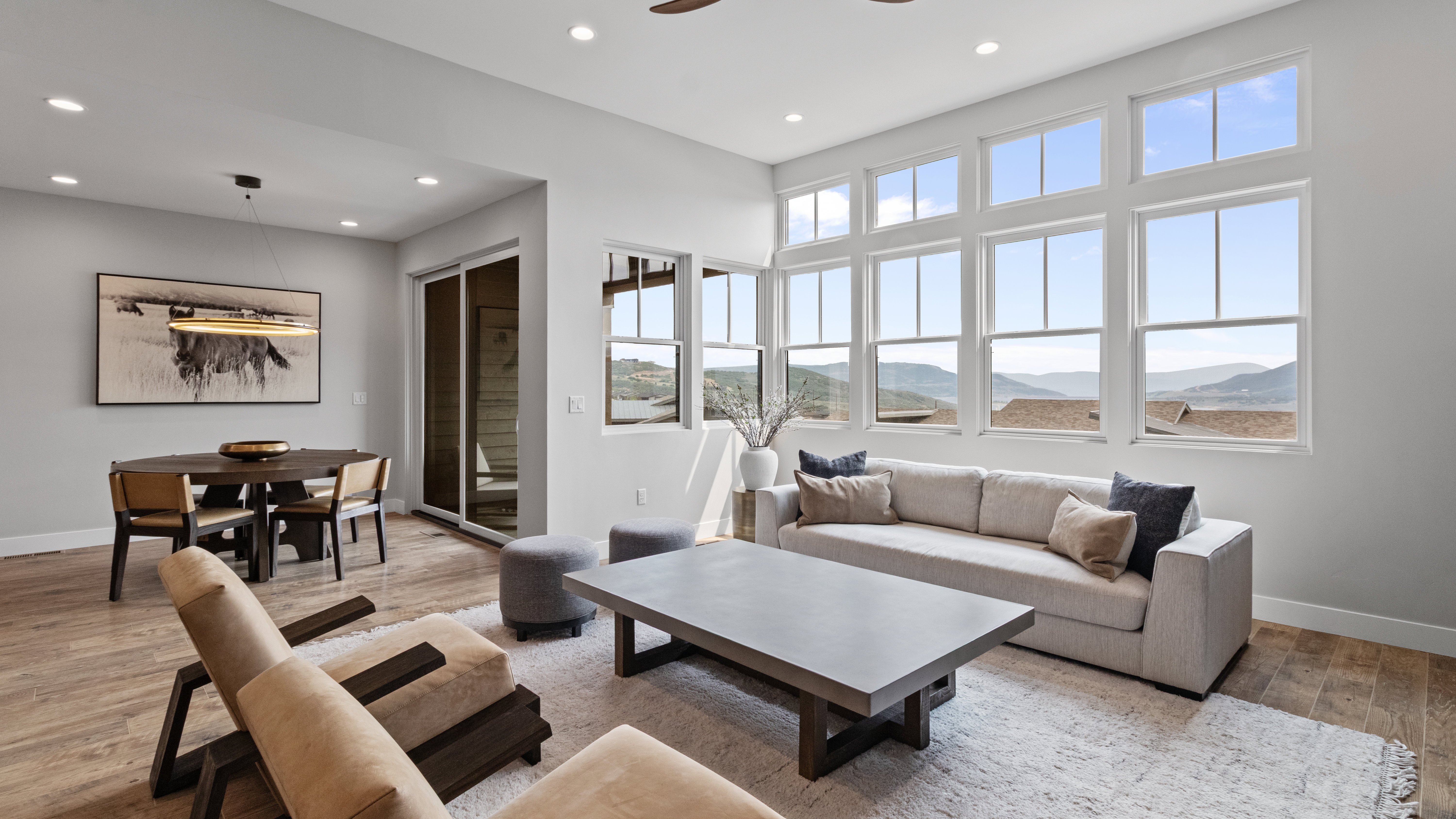 Expansive living area featuring a wall of multi-pane windows, wood-finish flooring, recessed lighting, and a contemporary dining space with a linear pendant light