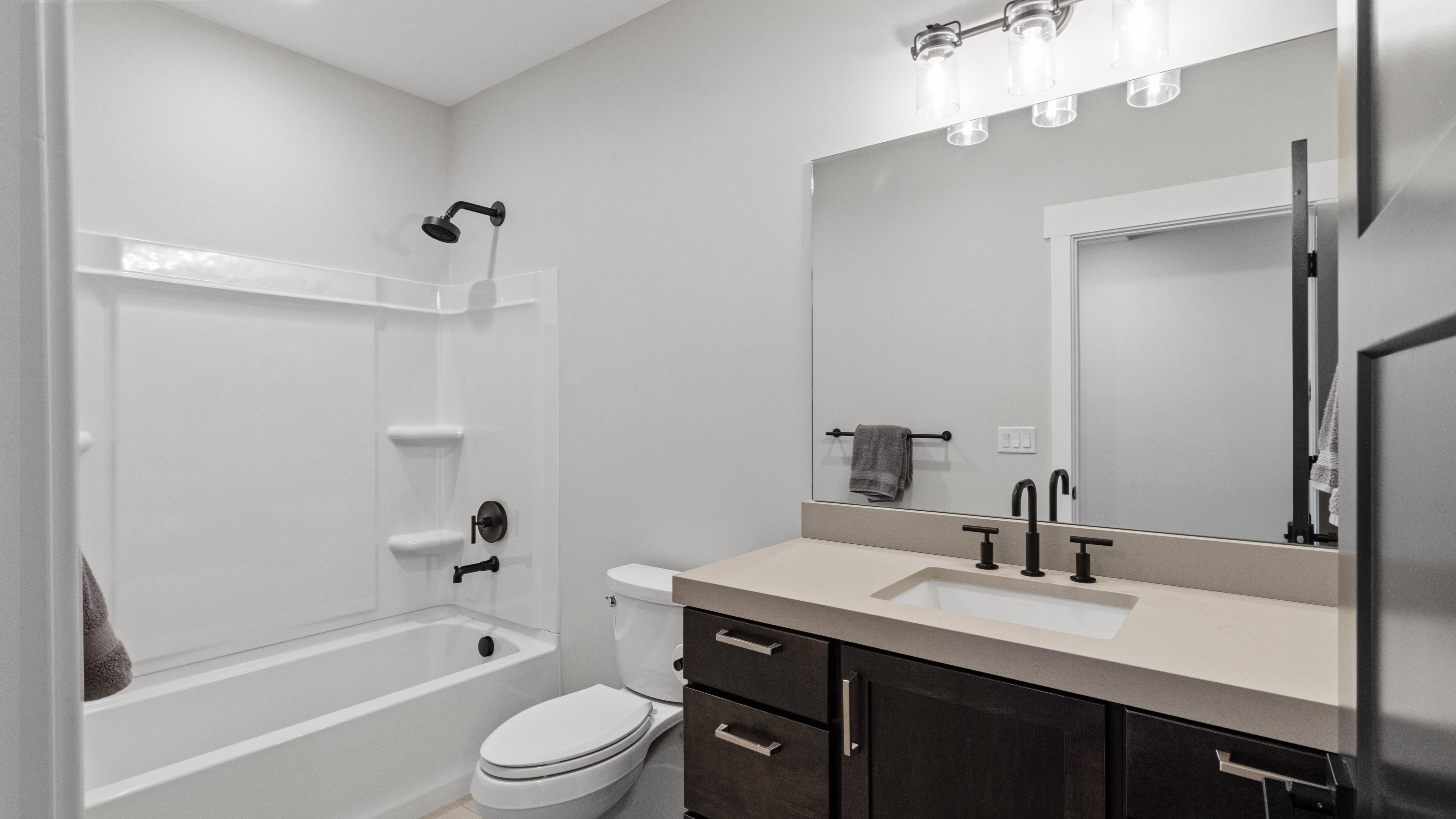 One of two lower level bathrooms featuring a white tub/shower combination with matte black fixtures, a white toilet, and a vanity with a light-colored countertop and dark wood-finish cabinetry
