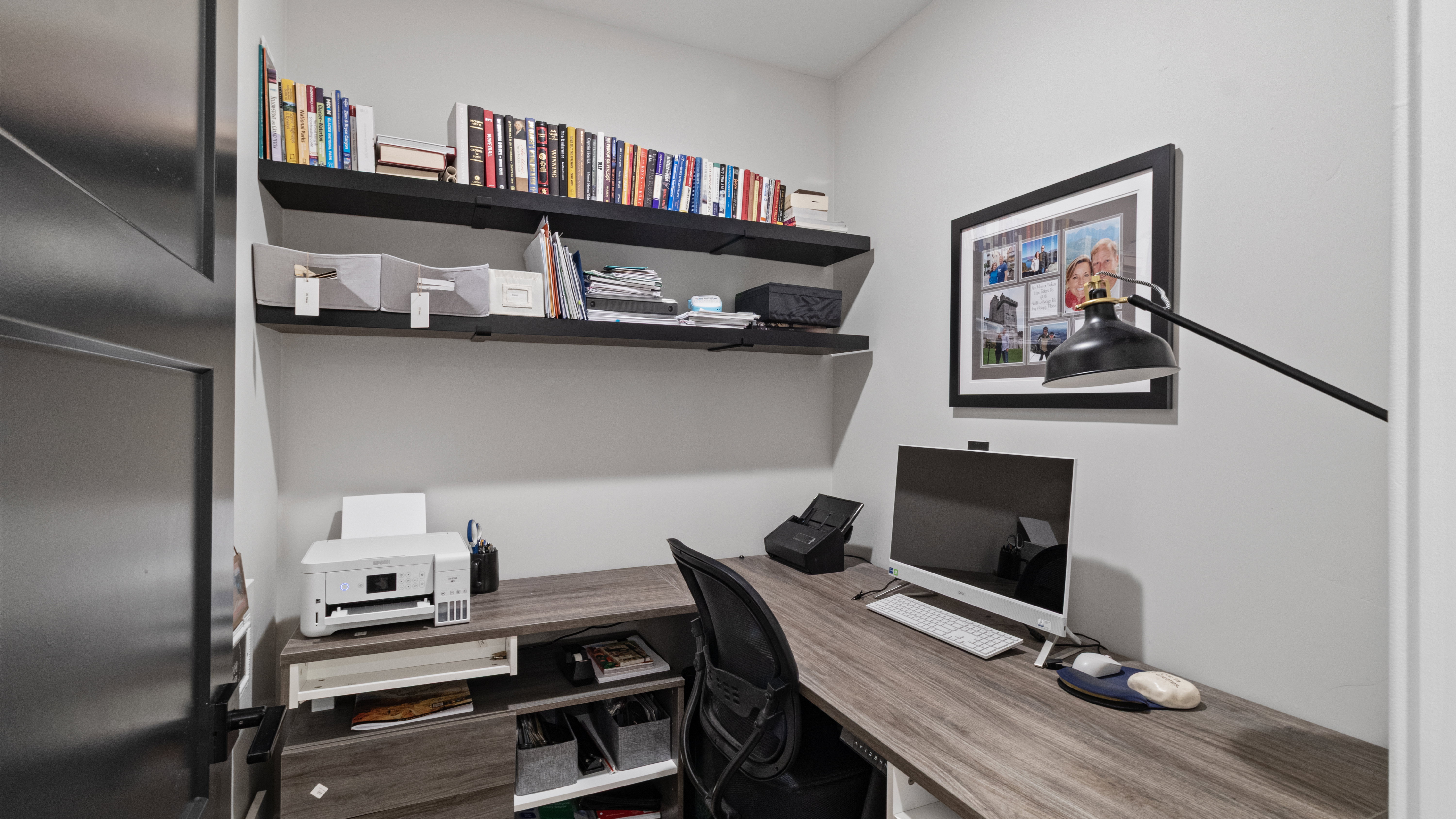 Pocket office with dedicated workspace featuring a wood-finish L-shaped desk with integrated shelving, wall-mounted floating shelves, and a contemporary matte black door