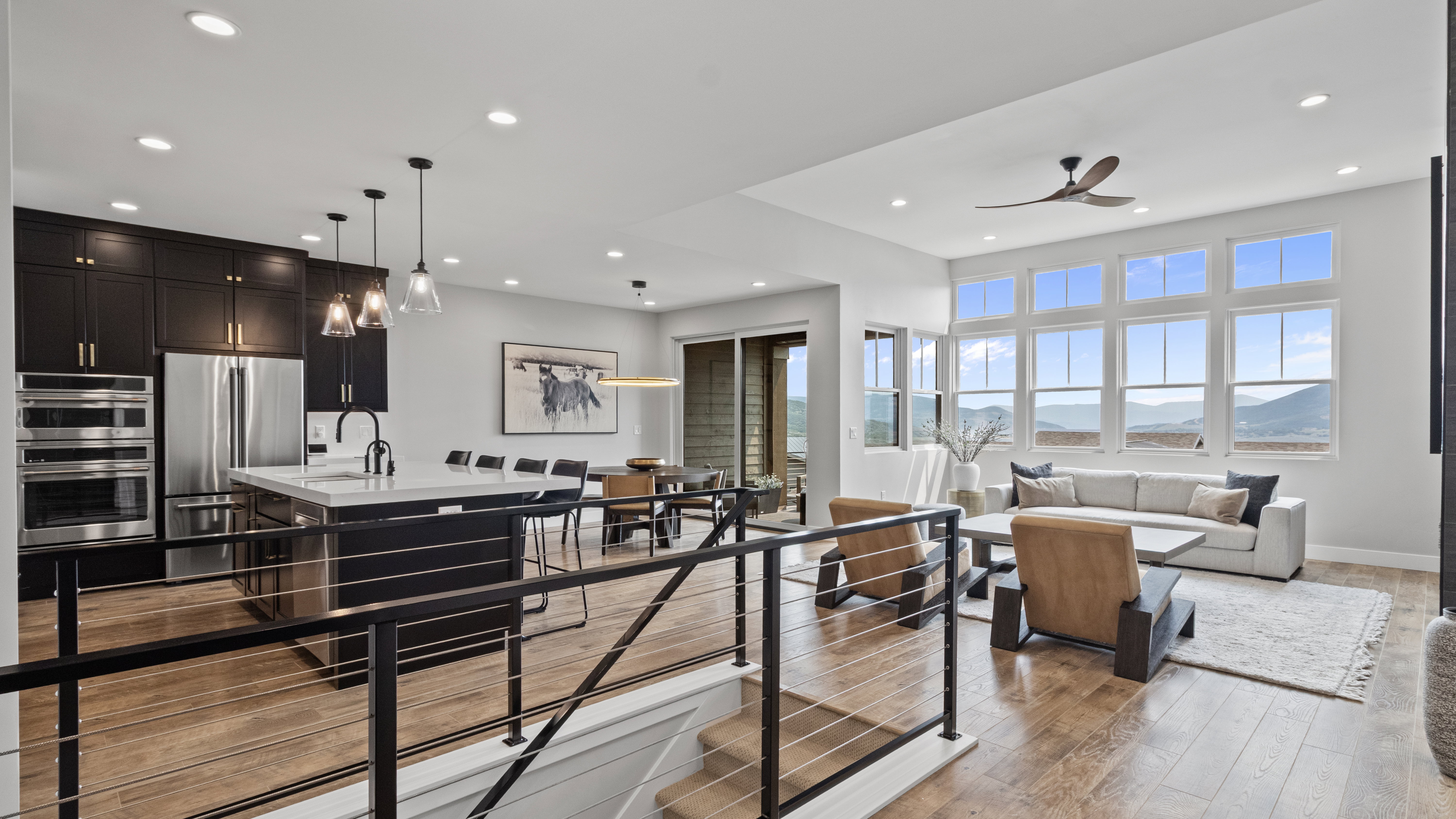 Open-concept living space with wood-finish flooring, a contemporary kitchen featuring dark cabinetry and stainless steel appliances, a center island with seating, a dining area, and a living room with a prominent wall of windows
